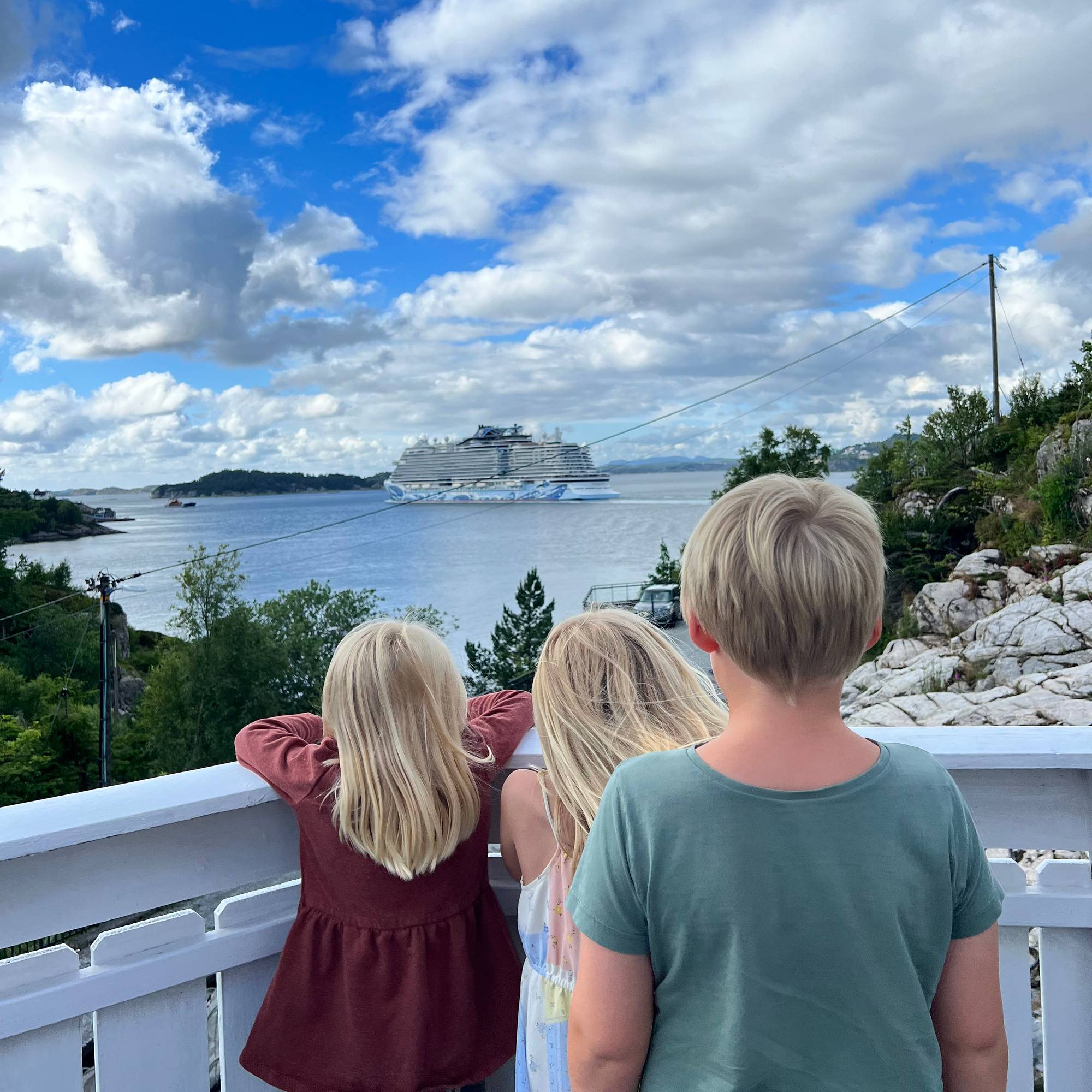 Tre blondebarn, to jenter og ein gut, står på ein altan ved eit rekkverk og kikar utover mot sjøen og eit cruiseskip i det fjerne.