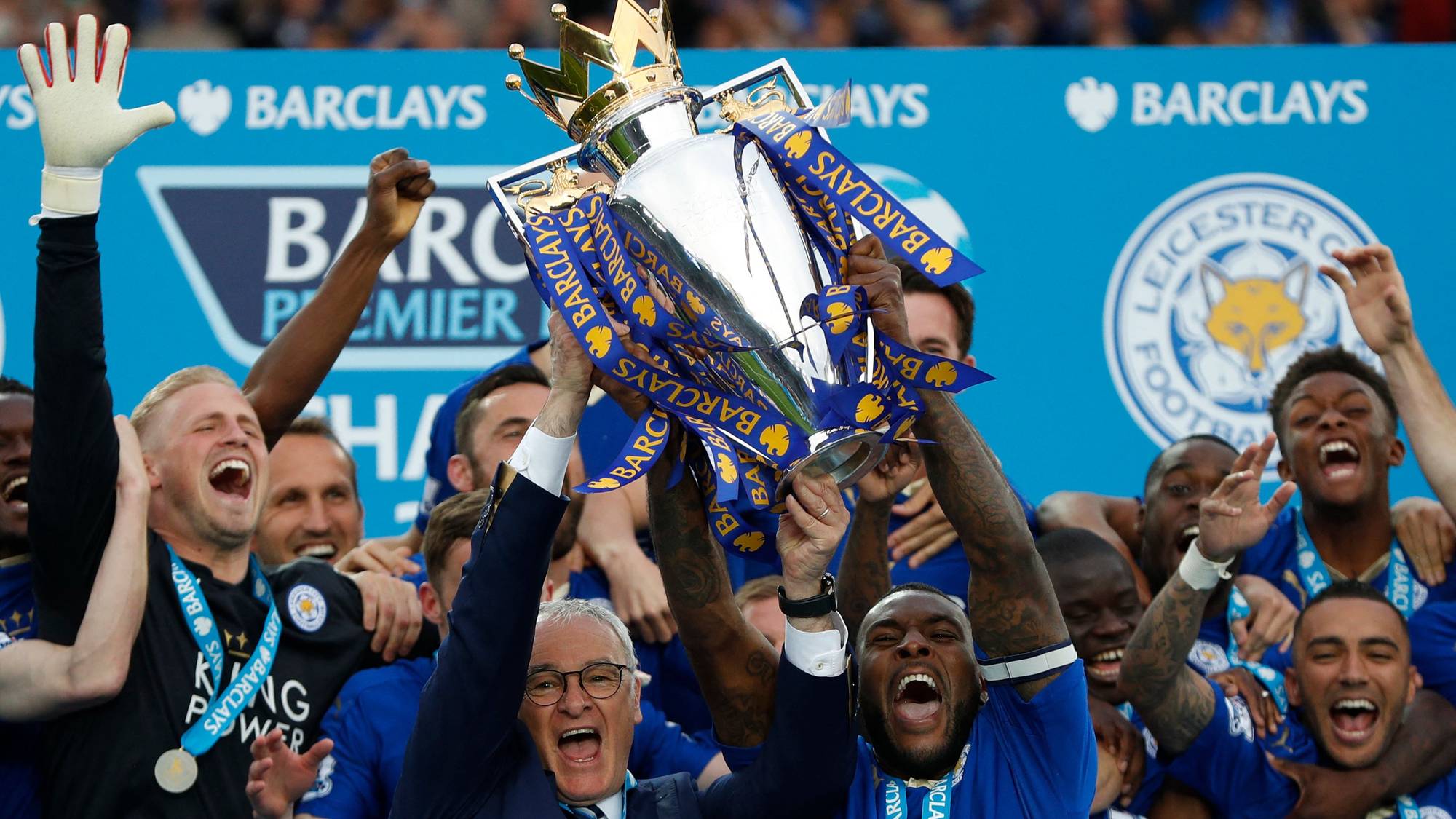 Leicester City-spillere feirer med Premier League-trofeet. Laget vant tittelen i 2016. En stor gruppe spillere er samlet rundt trofeet. Bak dem er det et banner med "Barclays Premier League". (Bildebeskrivelsen er laget av en KI-tjeneste)
