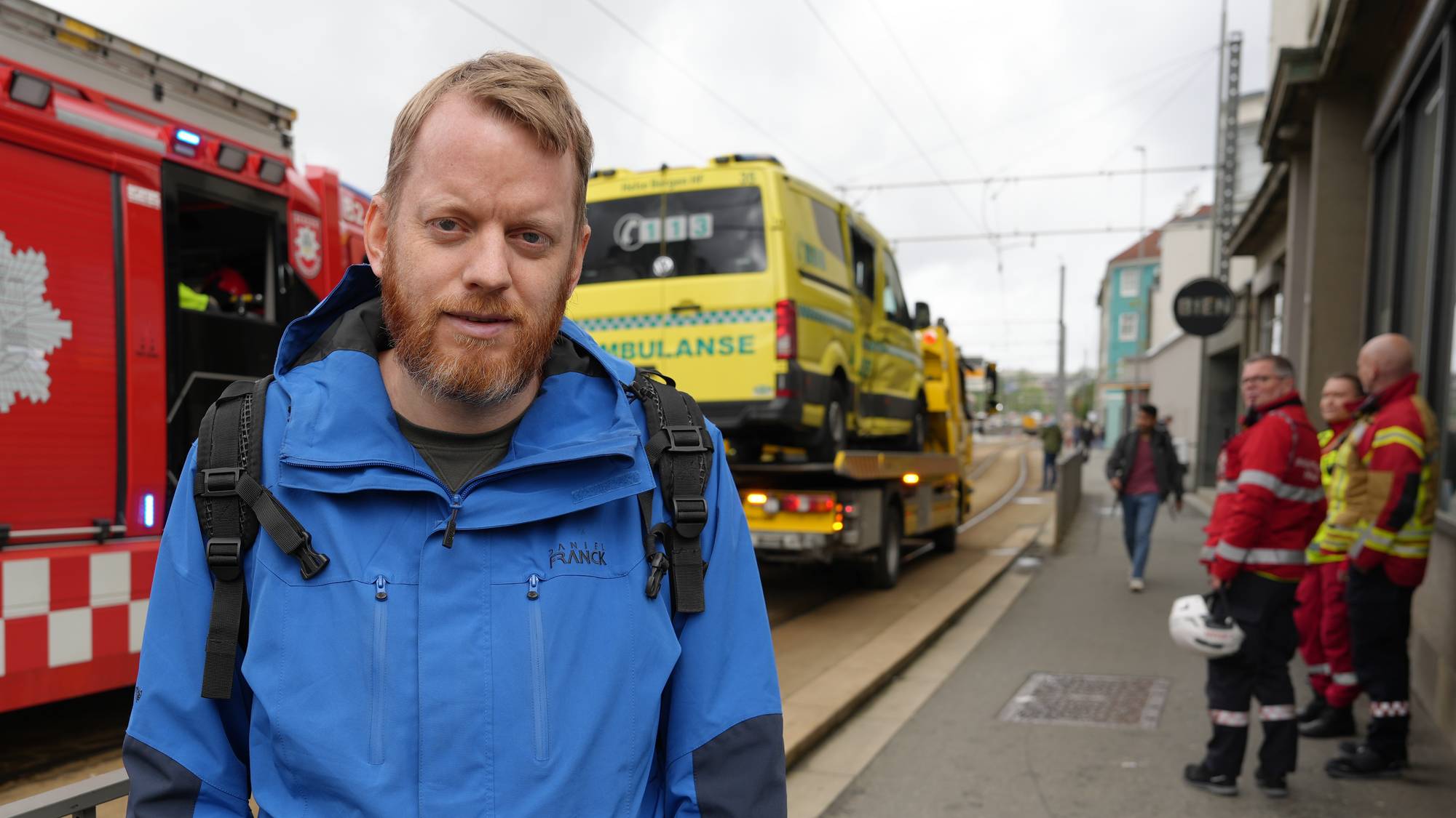 ein mann med jakke og sekk står framfor ein brannbil og ein ambulanse
