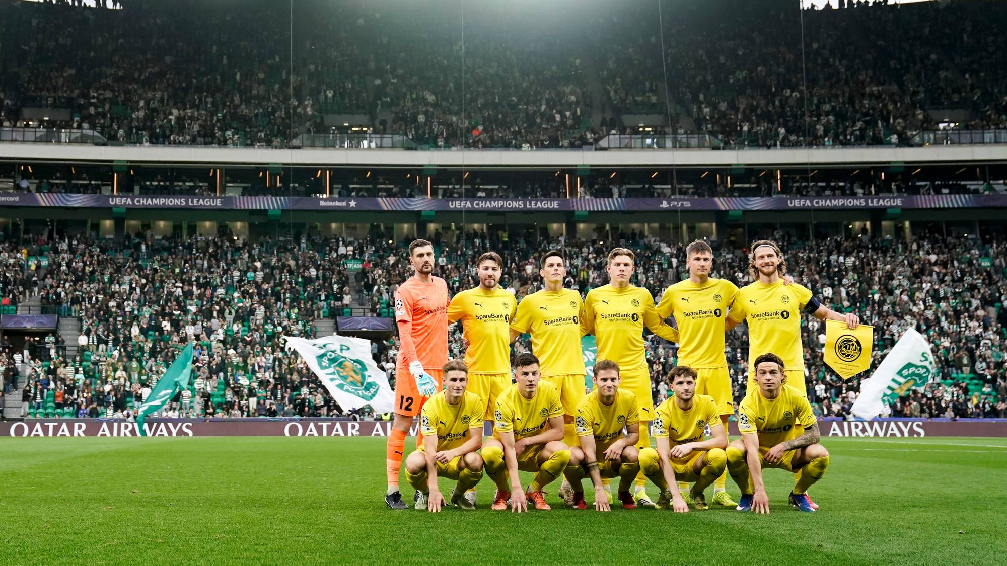 Et fotballag i gule drakter samler seg til lagbilde på en stadion med mange tilskuere. De sitter på gresset foran mål, mens goalkeeper står bak. Flagg med klubbens logo heves av spillerne. Stemningen virker energisk med en tydelig supporterbakgrunn. (Bildebeskrivelsen er laget av en KI-tjeneste)