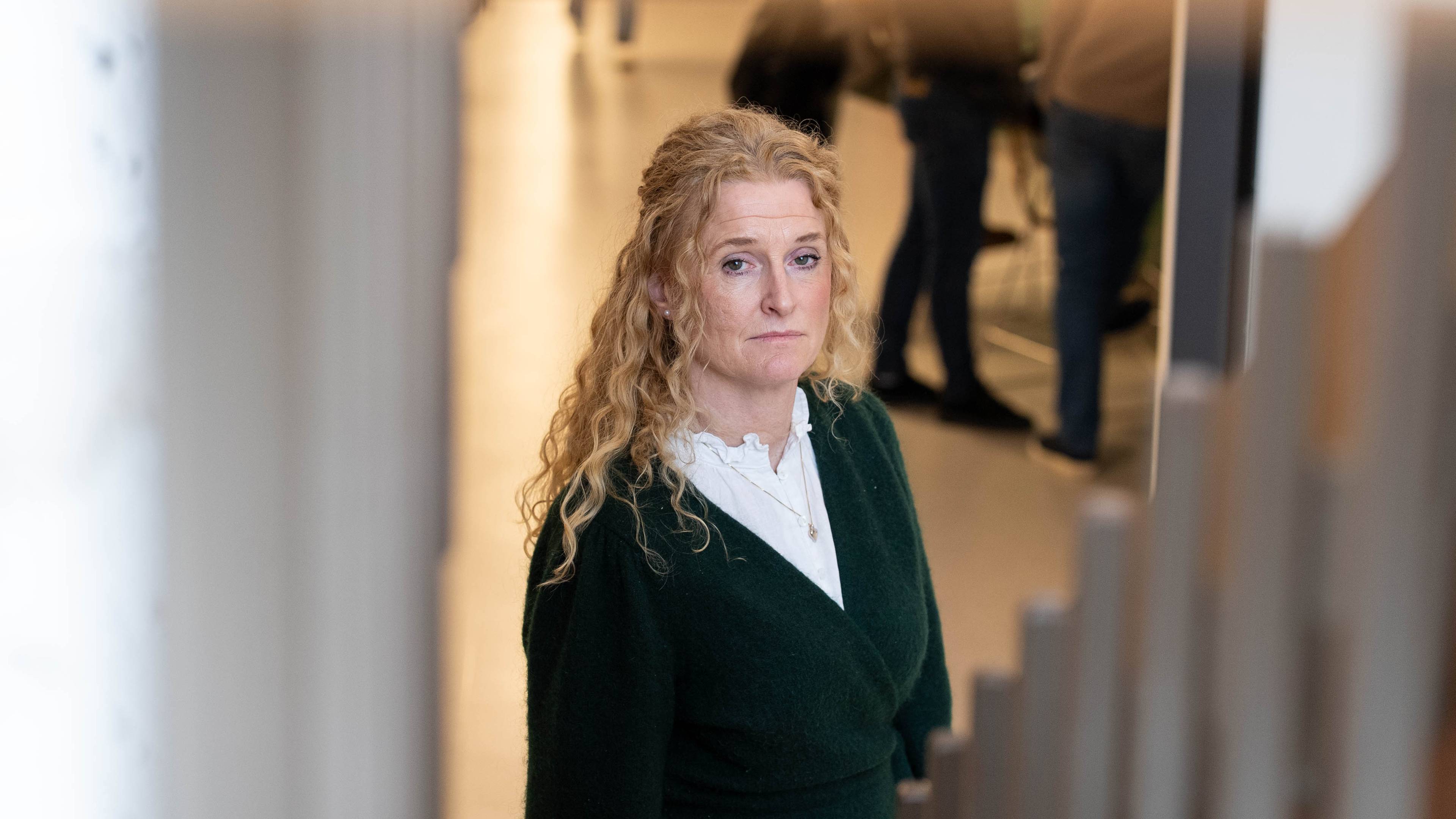 En kvinne står i en trapp og ser mot kameraet. Hun har blondt, krøllete hår og er kledd i en mørkegrønn genser over en hvit bluse. Bak henne er andre personer i uskarpt bakgrunnslys. (Bildebeskrivelsen er laget av en KI-tjeneste)