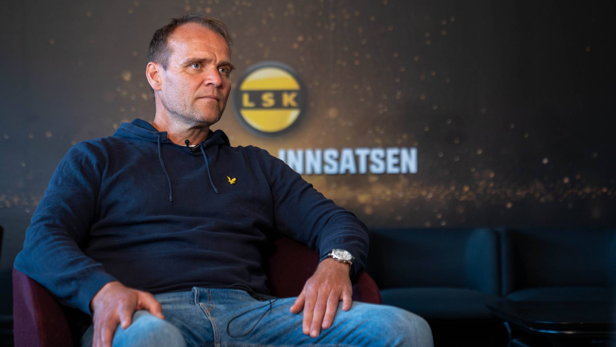 En mann sitter på en mørk sofa foran en vegg med ovale portrettbilder. Bildene viser ansiktene til flere personer i rammer. Bakgrunnen er en lyseblå vegg.