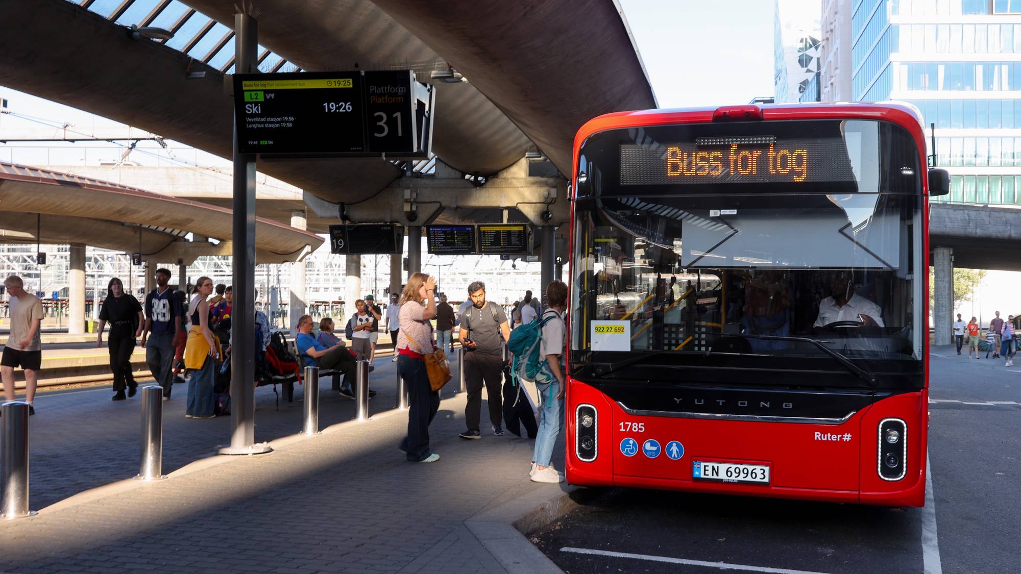Passasjerer venter ved en buss merket Buss for tog på en terminal ved et jernbanespor. (Bildebeskrivelsen er laget av en KI-tjeneste)