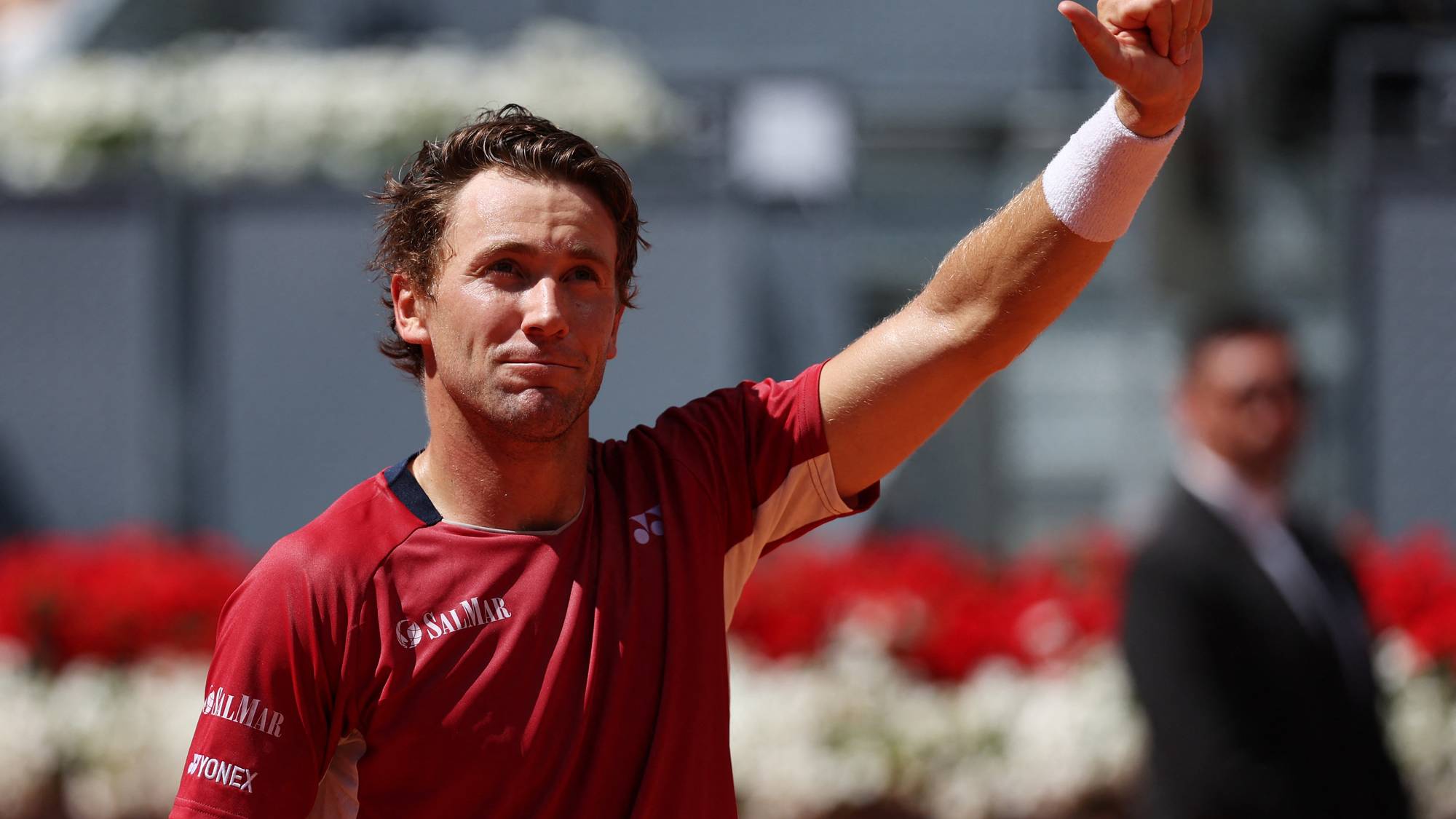 En tennisspiller med en rød trøye som har reklame på, løfter armen. Han har et svett pannebånd på armen. I bakgrunnen sitter tilskuere. (Bildebeskrivelsen er laget av en KI-tjeneste)