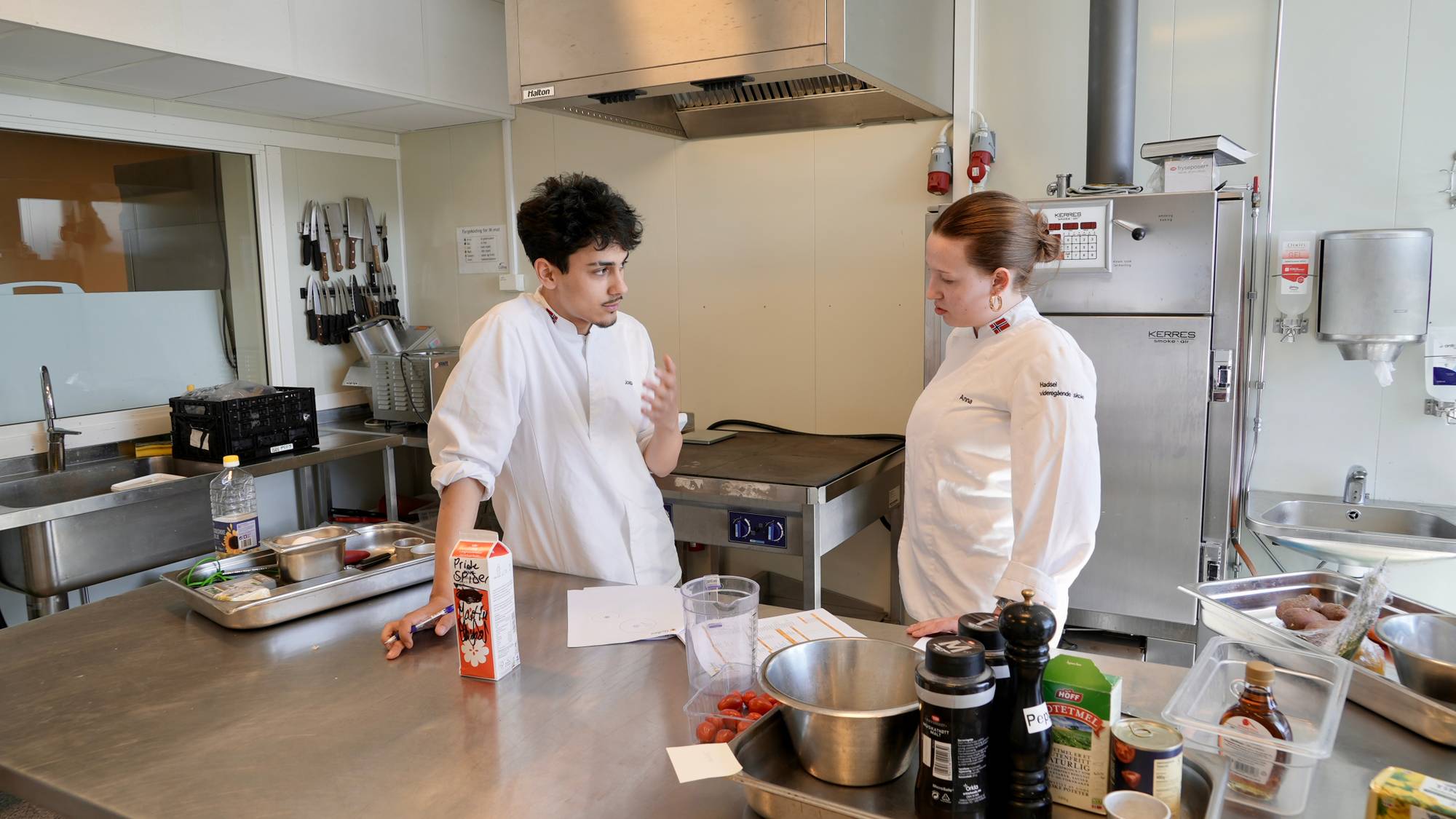 To kokker i hvite uniformer diskuterer mens de står ved en benk på et kjøkken. Mannen gestikulerer mens kvinnen lytter. På benken foran dem står det ingredienser, blant annet en melkekartong og cherrytomater. En stor kjøleskap står til høyre i bildet. (Bildebeskrivelsen er laget av en KI-tjeneste)