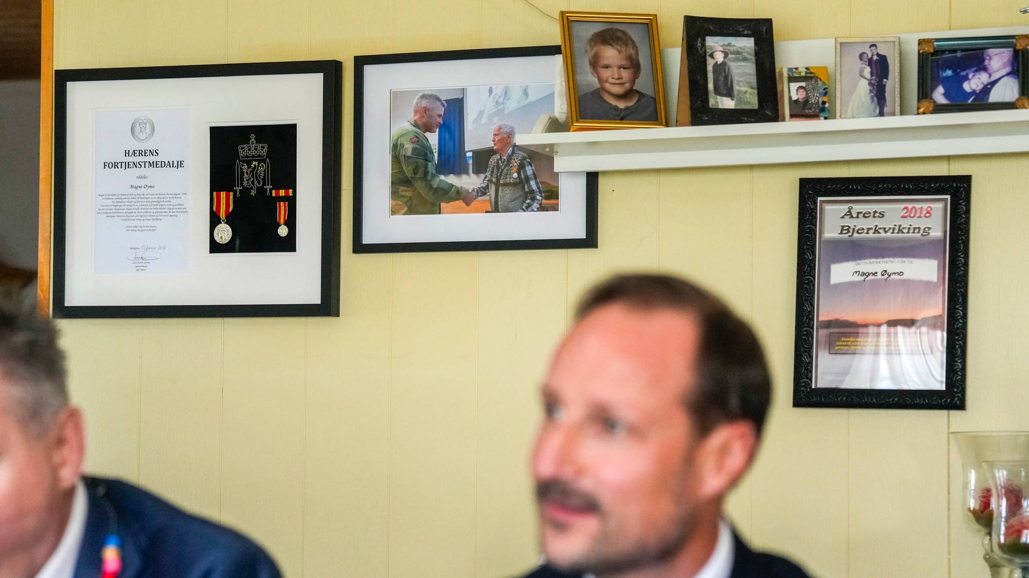 Kronprins Haakon sitter foran en vegg med innrammede bilder og diplomer i et rom. (Bildebeskrivelsen er laget av en KI-tjeneste)