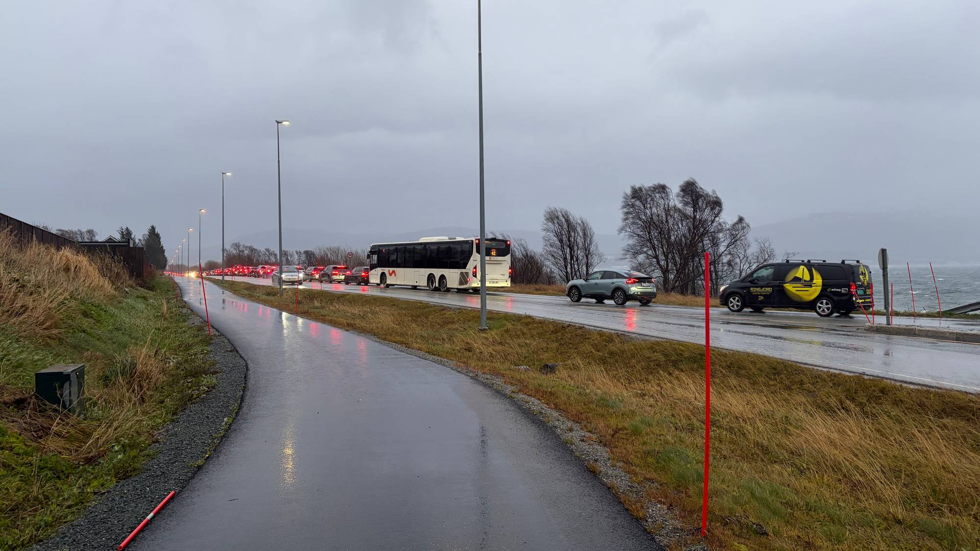 Kø med biler og en buss på en våt vei langs sjøen under grå himmel. (Bildebeskrivelsen er laget av en KI-tjeneste)
