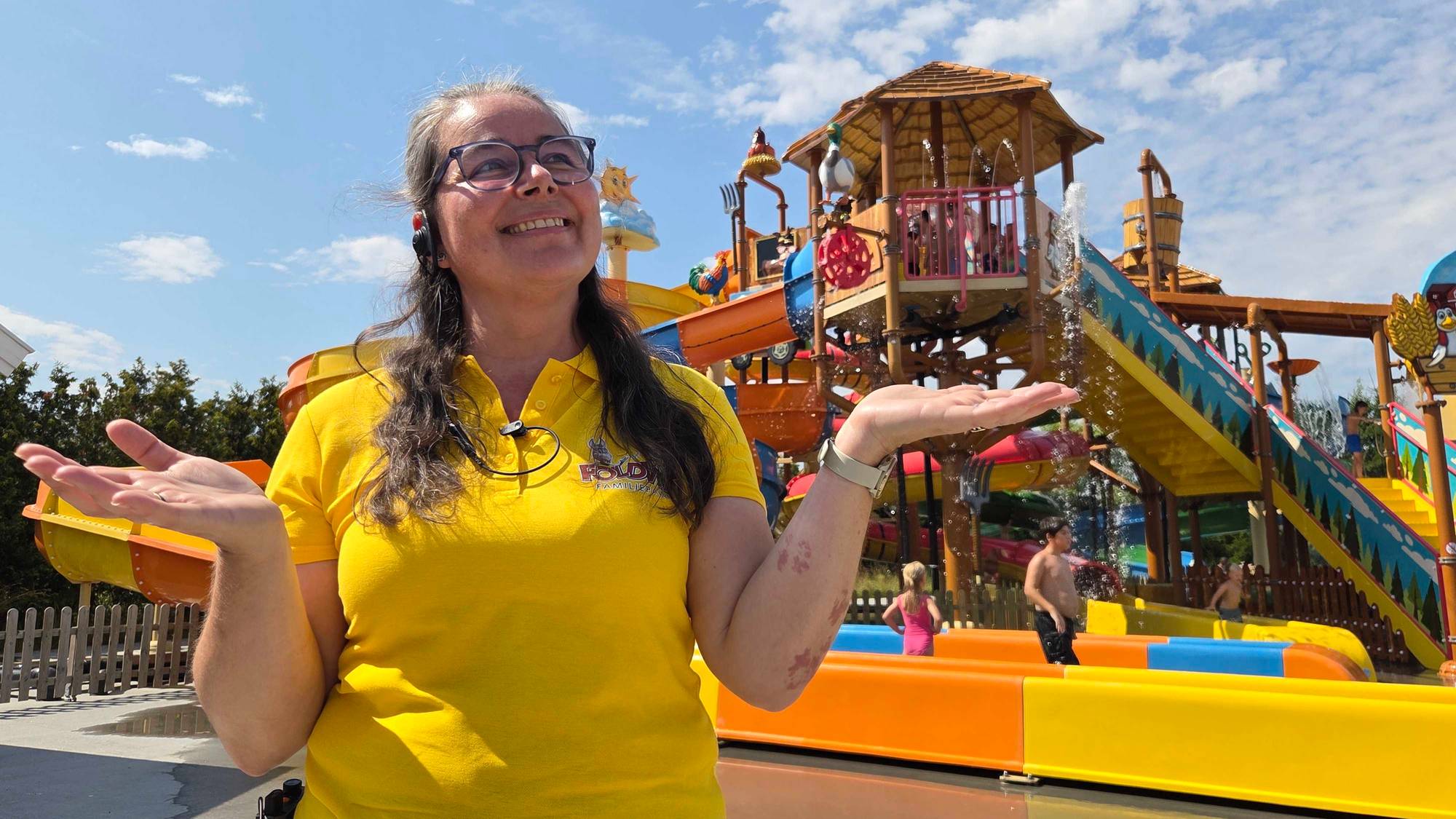 kvinne med langt hår og briller iført gul tksjorte smiler til kamera. familiepark i bakgrunnen