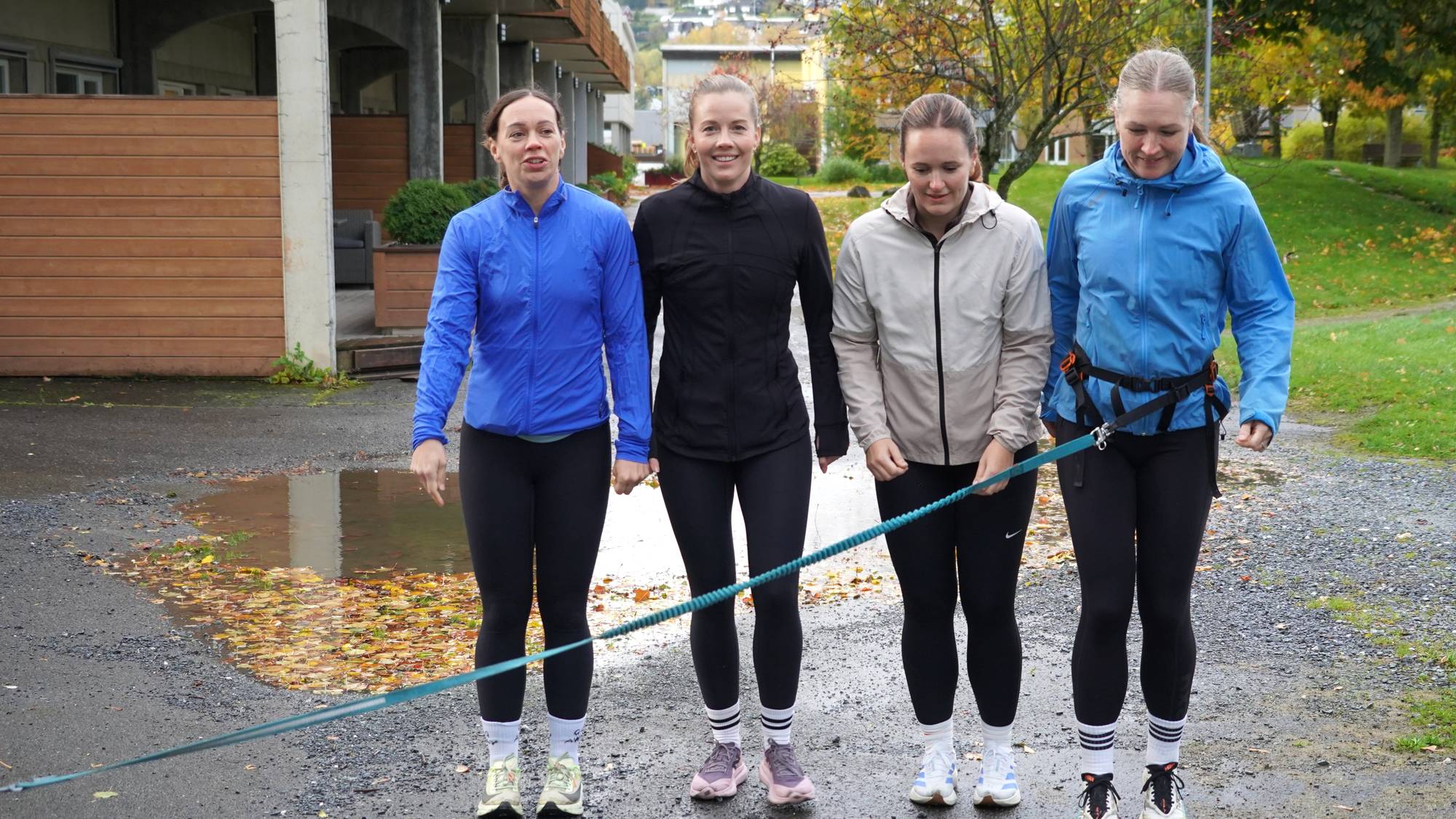 Fire damer står på rekke klare for å jogge. De er iført jakke og tights, og det er vått på bakken.