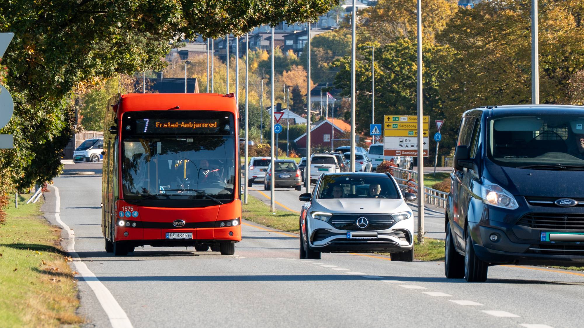 Rød buss og biler kjører på en vei i dagslys. Veiskilt viser retning mot Fredrikstad.