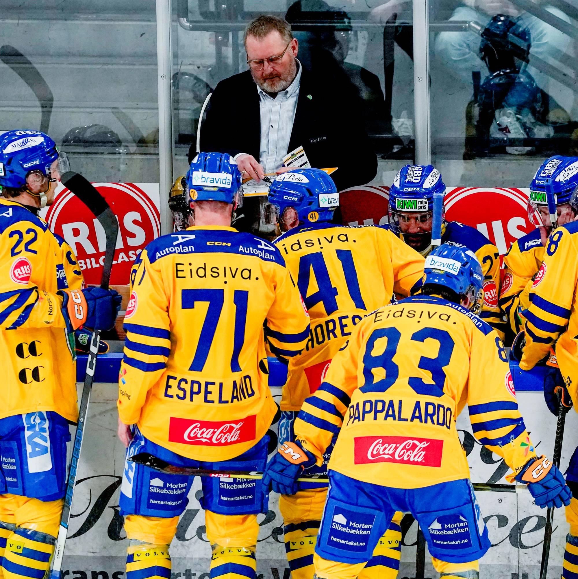 Et ishockeylag i gule og blå drakter står samlet med treneren bak. Spillere med nummer 71, 83 og 41 er synlige. De står på isen under en kamp. (Bildebeskrivelsen er laget av en KI-tjeneste)