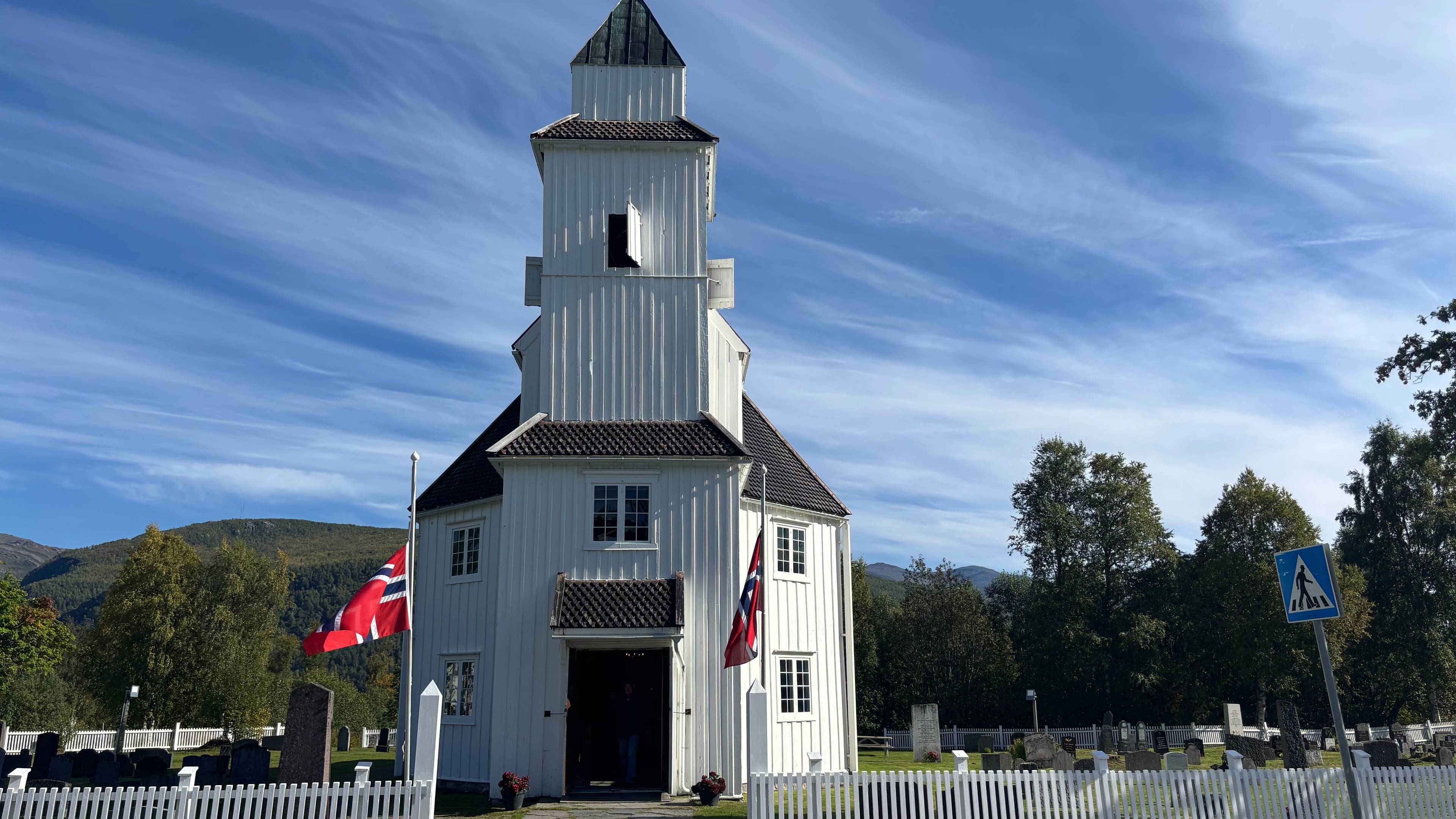 Hvit trekirke med to norske flagg og en kirkegård i forgrunnen under en blå himmel. (Bildebeskrivelsen er laget av en KI-tjeneste)