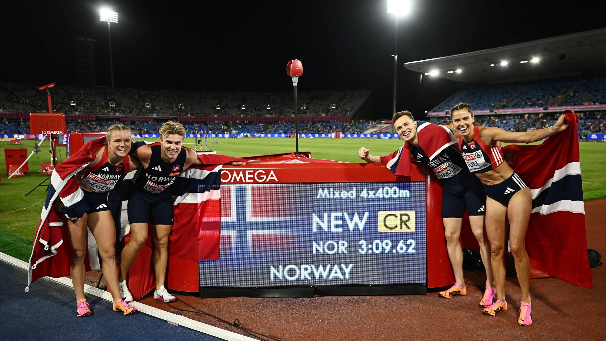 Fire norske utøvere feirer seier med norske flagg ved en resultattavle etter 4x400 meter stafett. (Bildebeskrivelsen er laget av en KI-tjeneste)