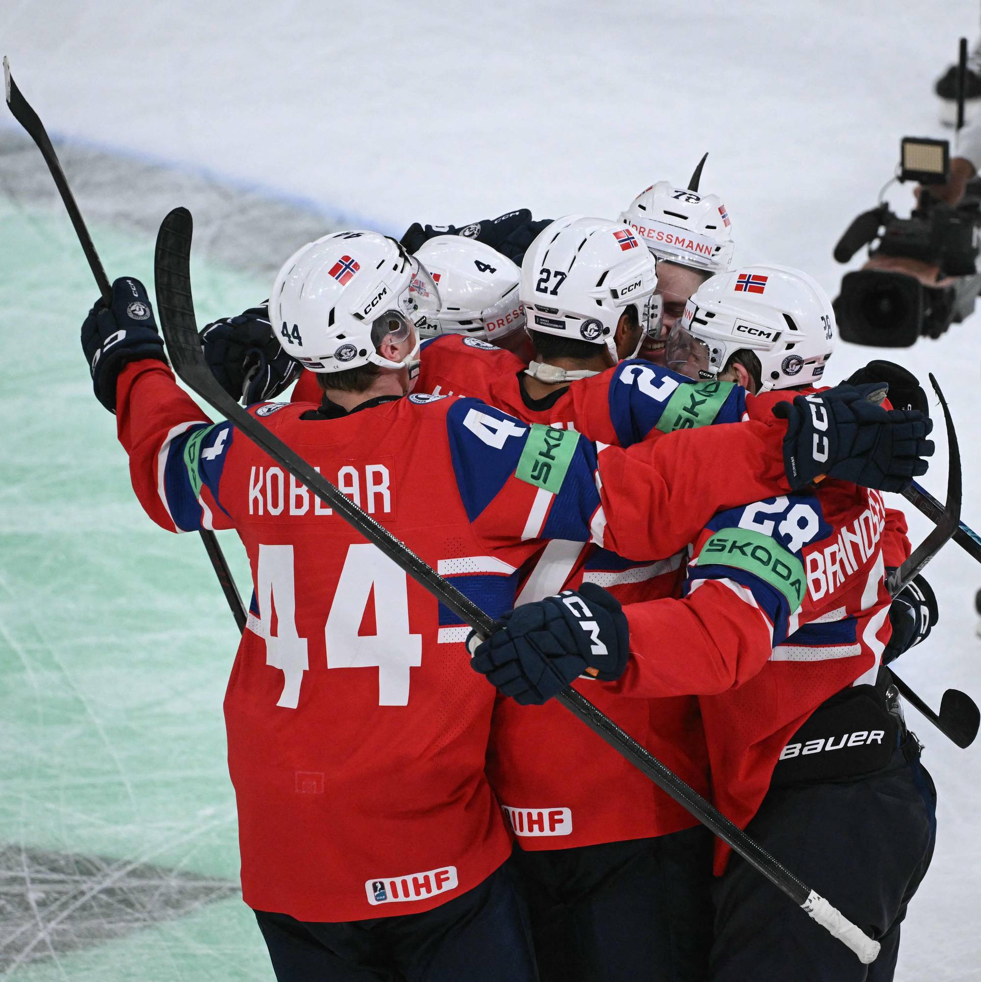 En gruppe ishockeyspillere feirer en scoring. Spillerne har på seg røde drakter med norske flagg på hjelmene. En TV-kameramann filmer i bakgrunnen. (Bildebeskrivelsen er laget av en KI-tjeneste)