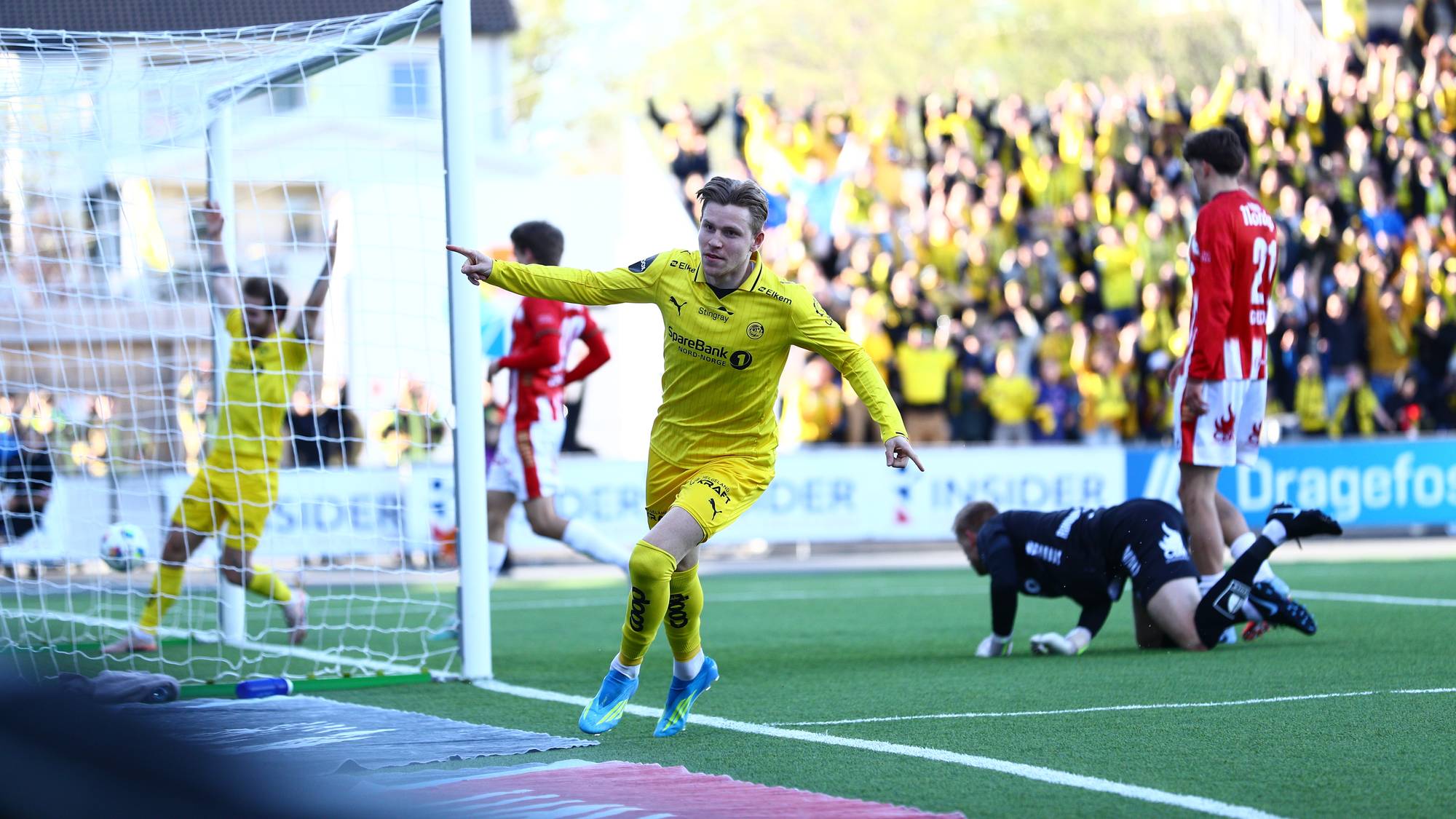 En fotballspiller i gult jubler etter å ha scoret mål. En keeper i svart ligger nede på gresset. I bakgrunnen sitter publikum på tribunen. (Bildebeskrivelsen er laget av en KI-tjeneste)
