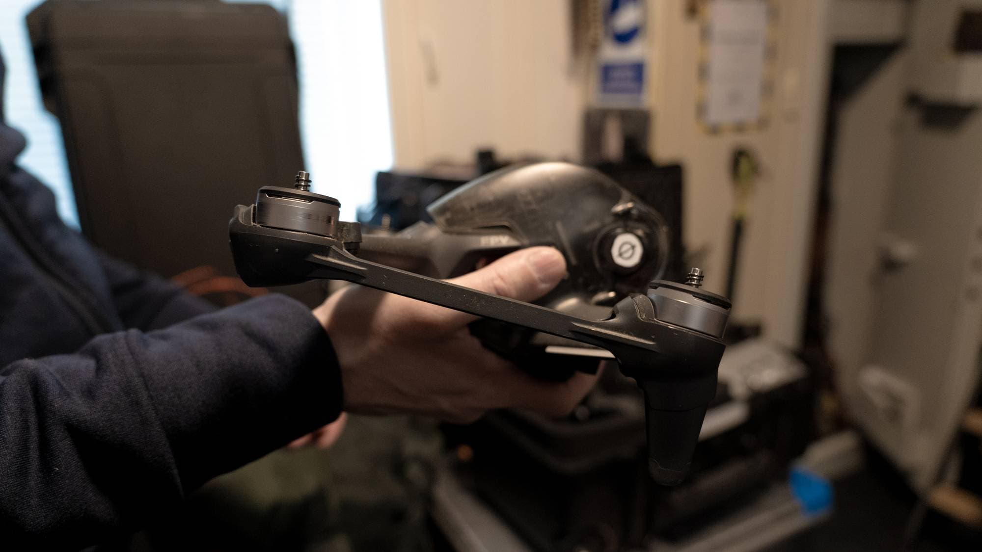 En person holder en drone i hånden. Dronen er svart med sølvfargede propellerfester. I bakgrunnen er det noe utstyr og et vindu. Det ser ut til å være innendørs. (Bildebeskrivelsen er laget av en KI-tjeneste)