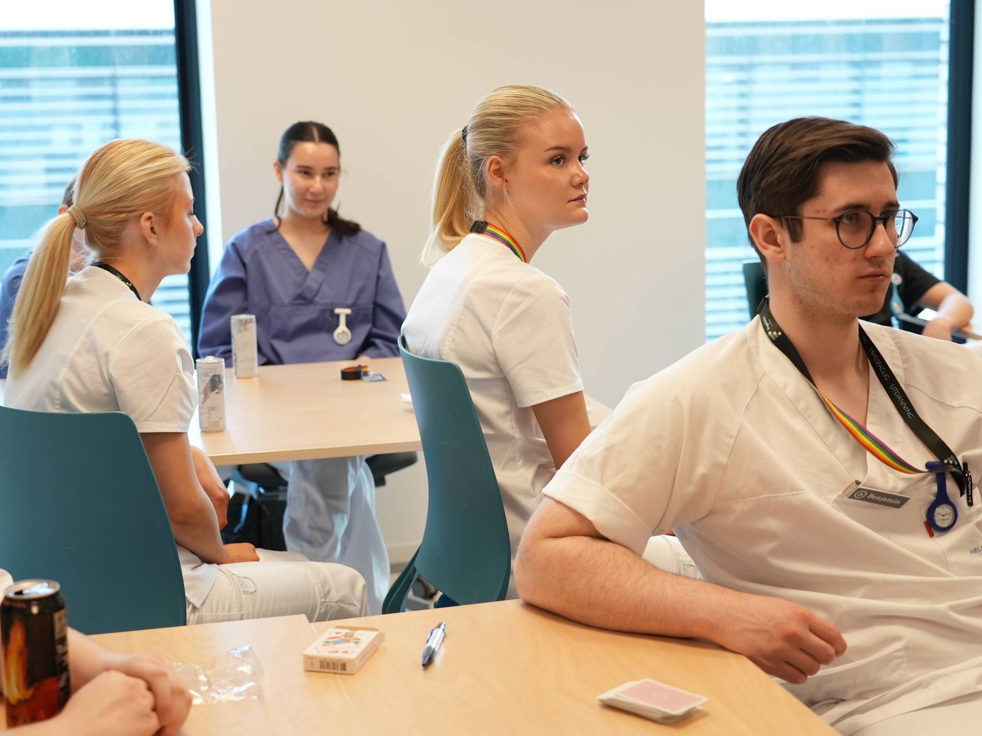 Flere studenter i hvite og blå uniformer sitter rundt et bord. En mann i hvitt, med briller og regnbuefarget snor, ser mot høyre. En kvinne med hestehale i hvit uniform ser mot venstre. En kvinne i blå uniform sitter i bakgrunnen. (Bildebeskrivelsen er laget av en KI-tjeneste)