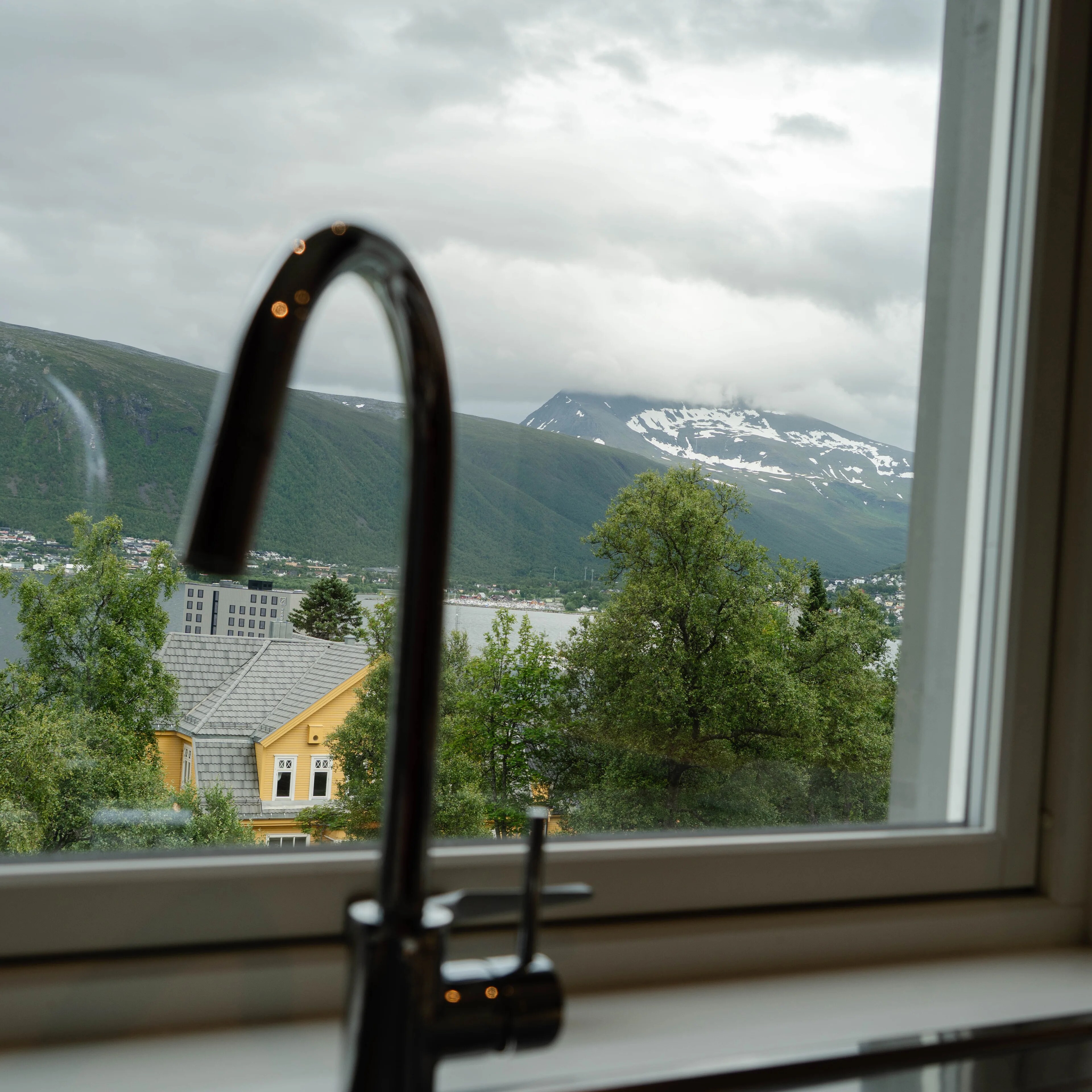 Utsikt fra et vindu med kjøkkenkran i forgrunnen. I bakgrunnen ses fjell, vann og bebyggelse. (Bildebeskrivelsen er laget av en KI-tjeneste)
