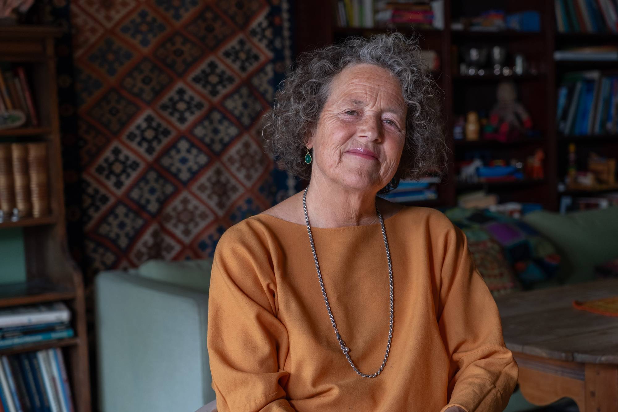 Eldre kvinne med grått krøllete hår i en oransje genser sitter foran en bokhylle og et teppe. 