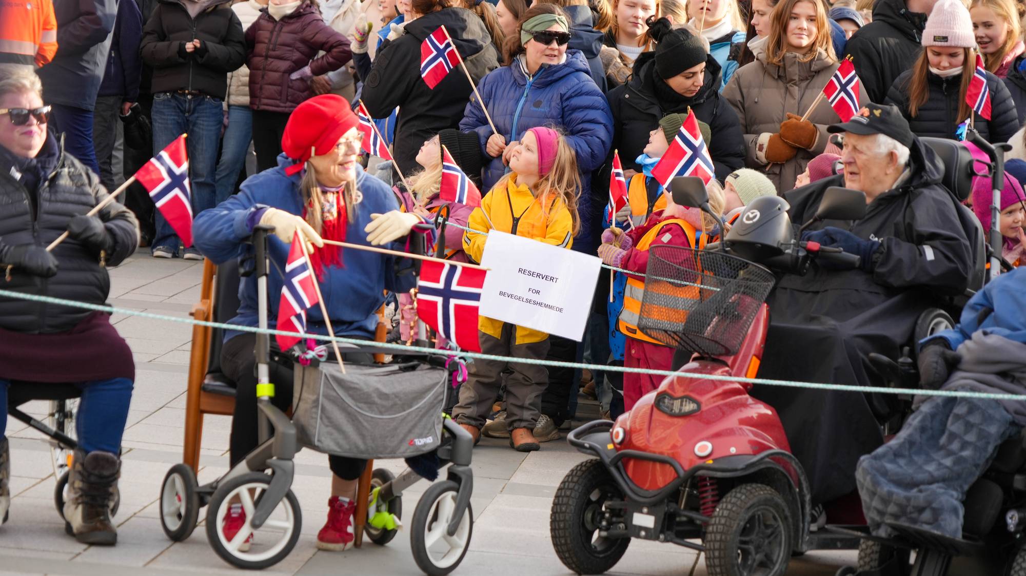 Gamle mennesker med rullator og gåstol venter på kongen. I bakgrunn står også barn med flagg.