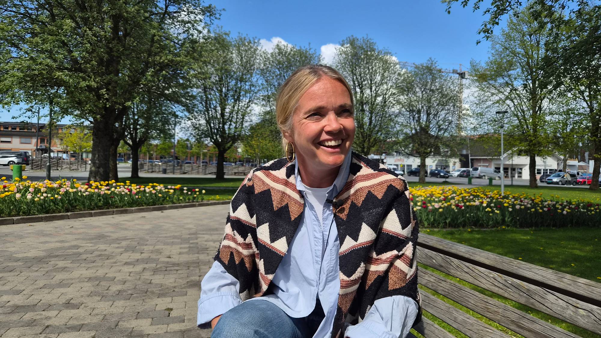 Kvinne smiler og ser til siden mens hun sitter på en parkbenk. Hun har på seg en lys skjorte, jeans og en fargerik vest. Bak henne er det et blomsterbed med tulipaner og trær. Solen skinner fra en blå himmel. (Bildebeskrivelsen er laget av en KI-tjeneste)