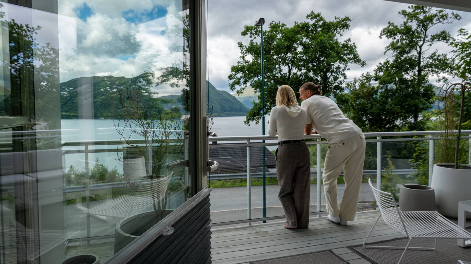 To personer står på en veranda og ser utover en fjord og fjellandskap. (Bildebeskrivelsen er laget av en KI-tjeneste)