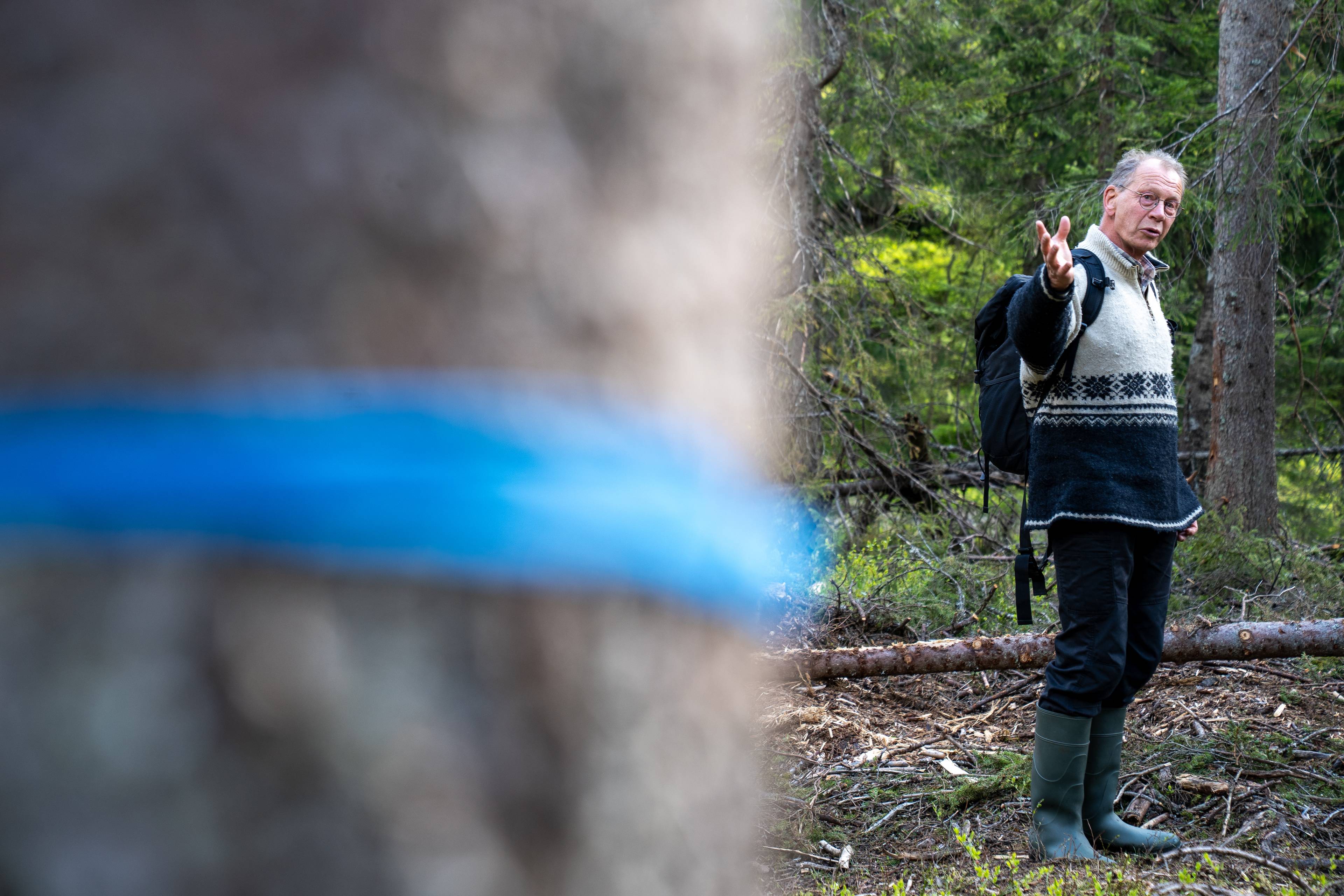 En mann i turklær og støvler står i skogen og gestikulerer. Han har en sekk på ryggen. Foran i bildet er en del av et tre med en blå stripe. (Bildebeskrivelsen er laget av en KI-tjeneste)