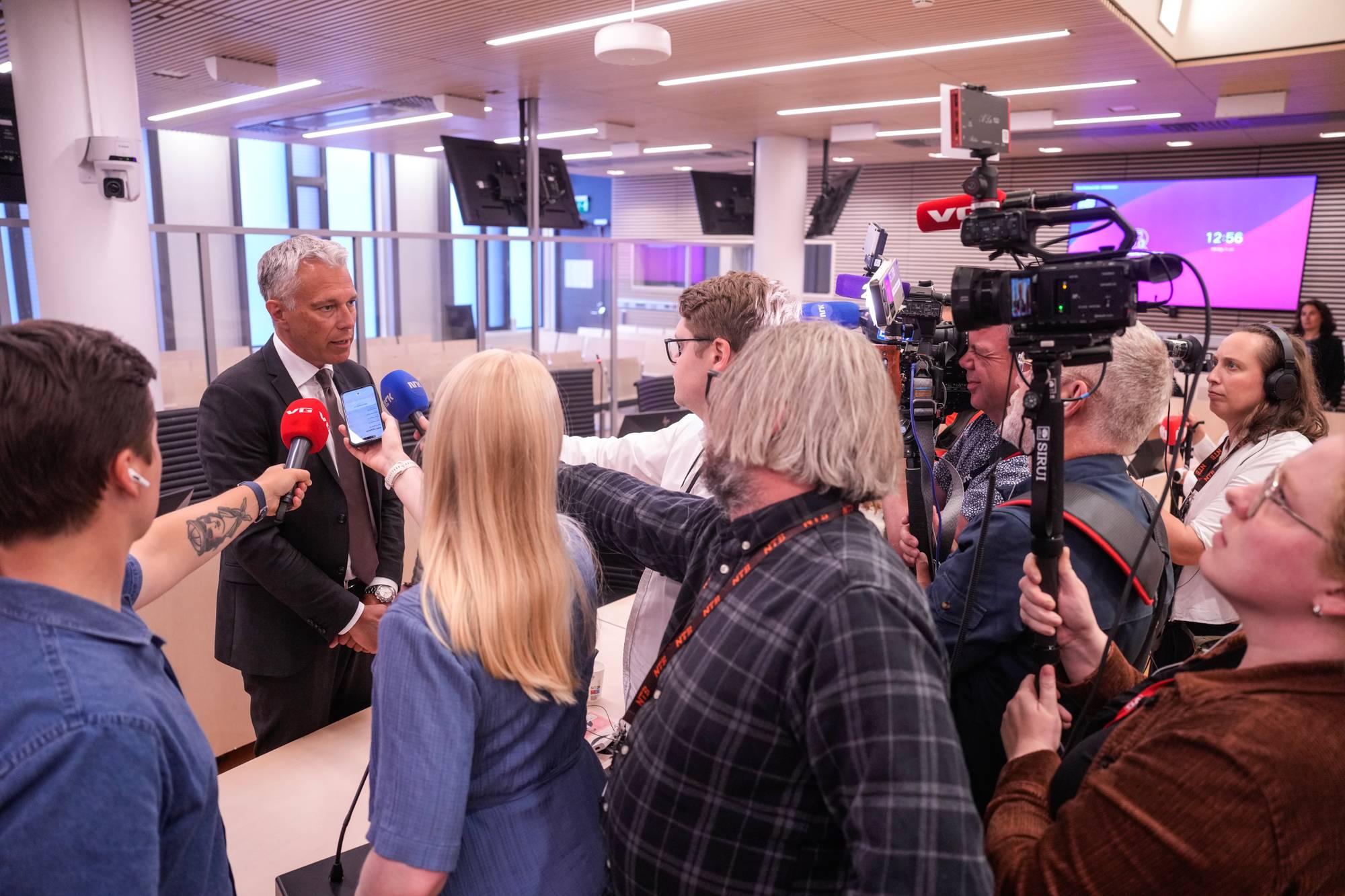 Mann i dress intervjues av en gruppe journalister med mikrofoner og kameraer i et kontorlandskap. (Bildebeskrivelsen er laget av en KI-tjeneste)