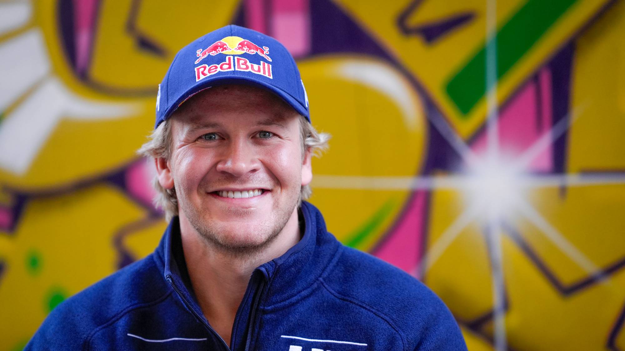 Aleksander Aamodt Kilde smiler foran en fargerik vegg med graffiti. (Bildebeskrivelsen er laget av en KI-tjeneste)