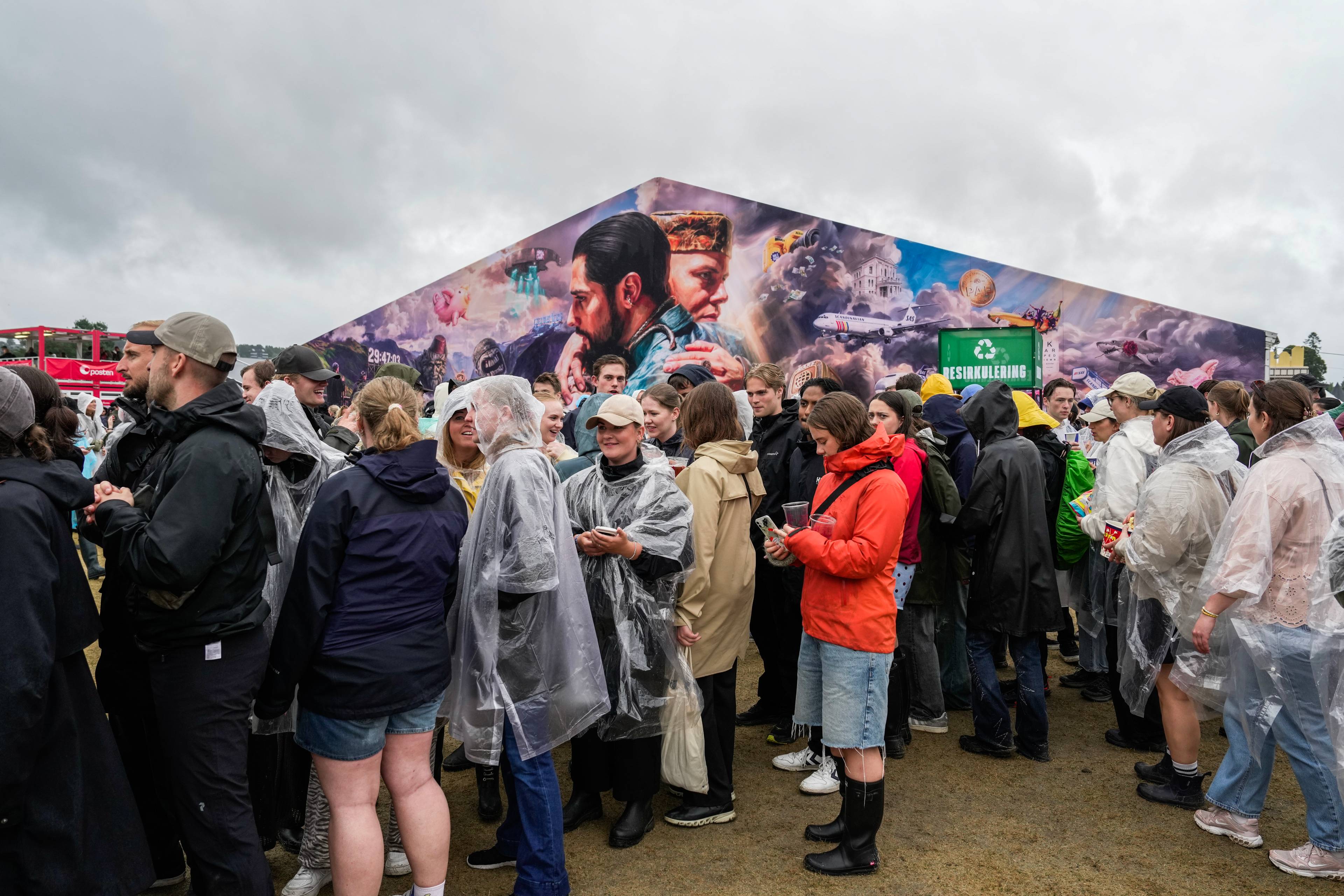 Publikum i regnklær står i kø ved et telt dekorert med et stort veggmaleri på en festival. (Bildebeskrivelsen er laget av en KI-tjeneste)