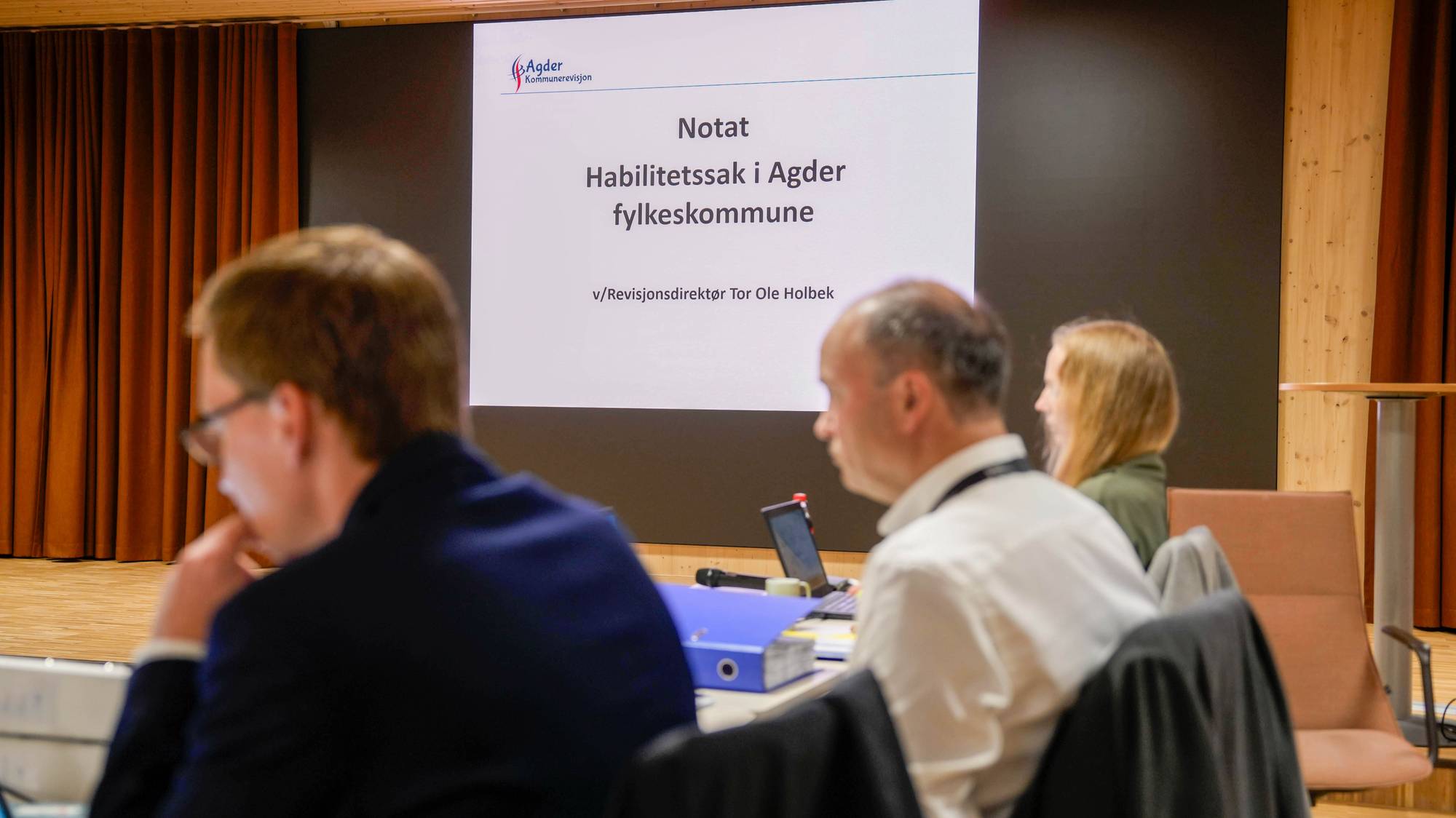 En skjerm viser en tittel om habilitetssak i Agder fylkeskommune. Tre personer sitter ved et bord foran skjermen, med ryggen til. De ser ut til å følge med på en presentasjon. Omvisning av et møterom med brune gardiner. (Bildebeskrivelsen er laget av en KI-tjeneste)