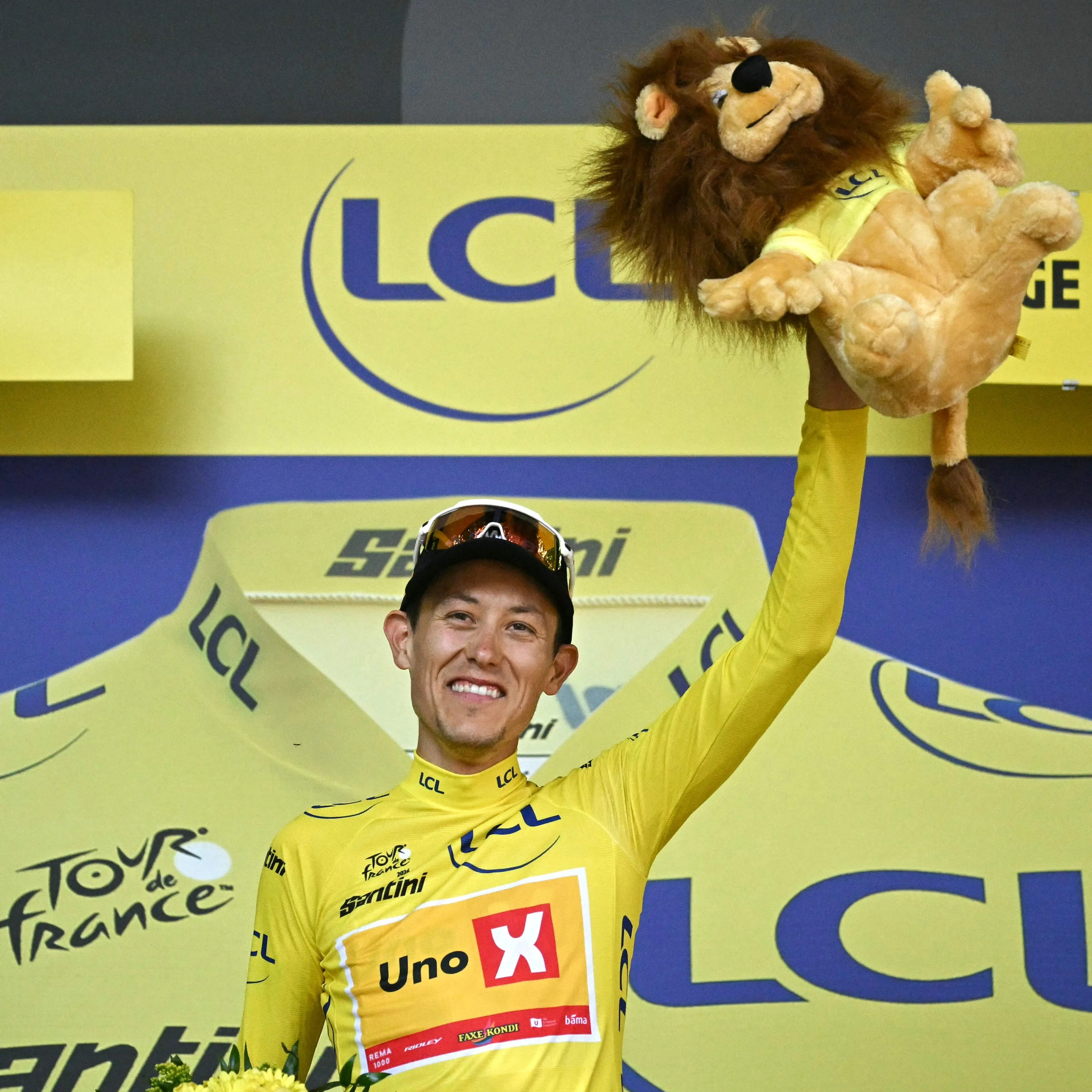 Syklist Jonas Abrahamsen i gul ledertrøye holder opp en kosedyr-løve på seierspallen i Tour de France. (Bildebeskrivelsen er laget av en KI-tjeneste)