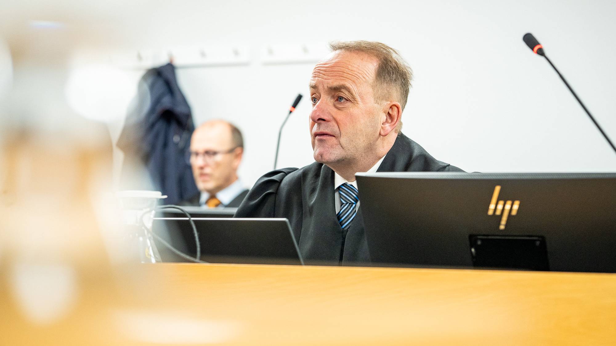 Bildet viser en domstolsscene med en dommer i kappe som sitter foran en dataskjerm. I bakgrunnen er en annen person delvis synlig. Det er mikrofoner plassert på bordet, og omgivelsene ser ut til å være et moderne rettslokale. Det er en enkel, nøytral atmosfære i rommet.
