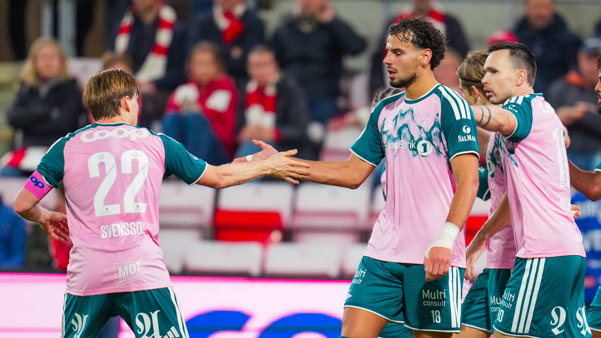 Fotballspillere i rosa og grønne drakter hilser på hverandre på banen foran publikum. (Bildebeskrivelsen er laget av en KI-tjeneste)
