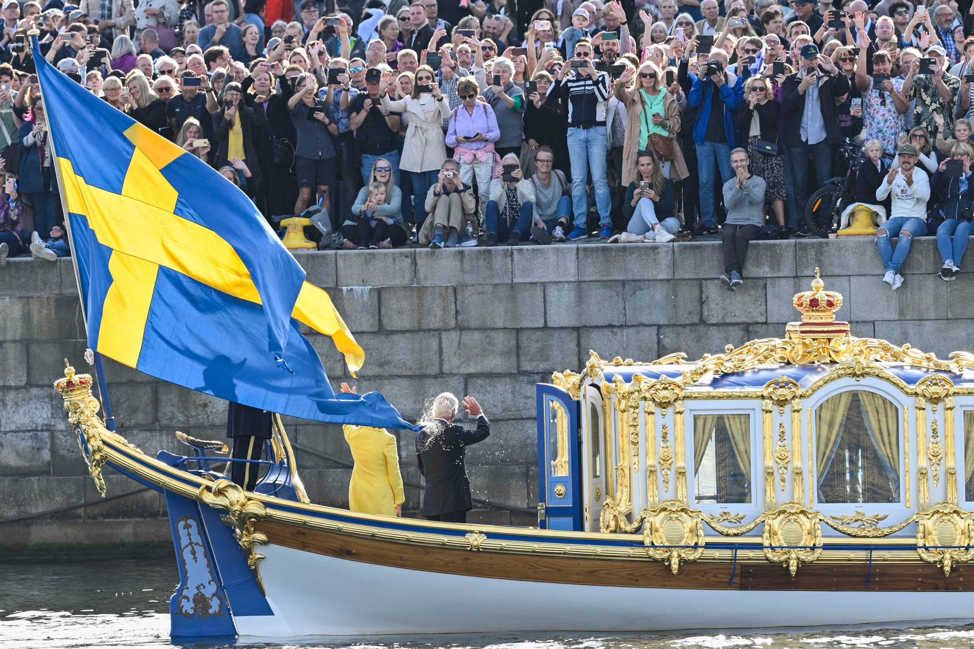 Kong Carl Gustaf og dronning Silvia vinker til folk som står langs kaia i Stockholm