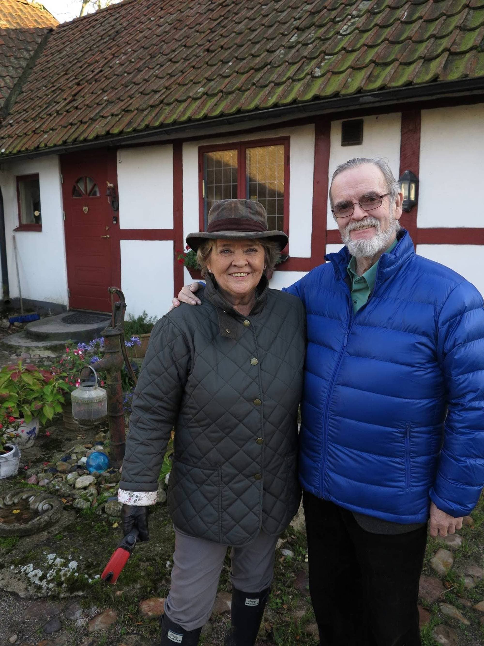 En eldre mann i blå boblejakke med solbrller står ved siden av en eldre kvinne i hatt og mørk jakke. De snakker sammen utendørs i det som ser ut som en kjøkkenhage utendørs. 