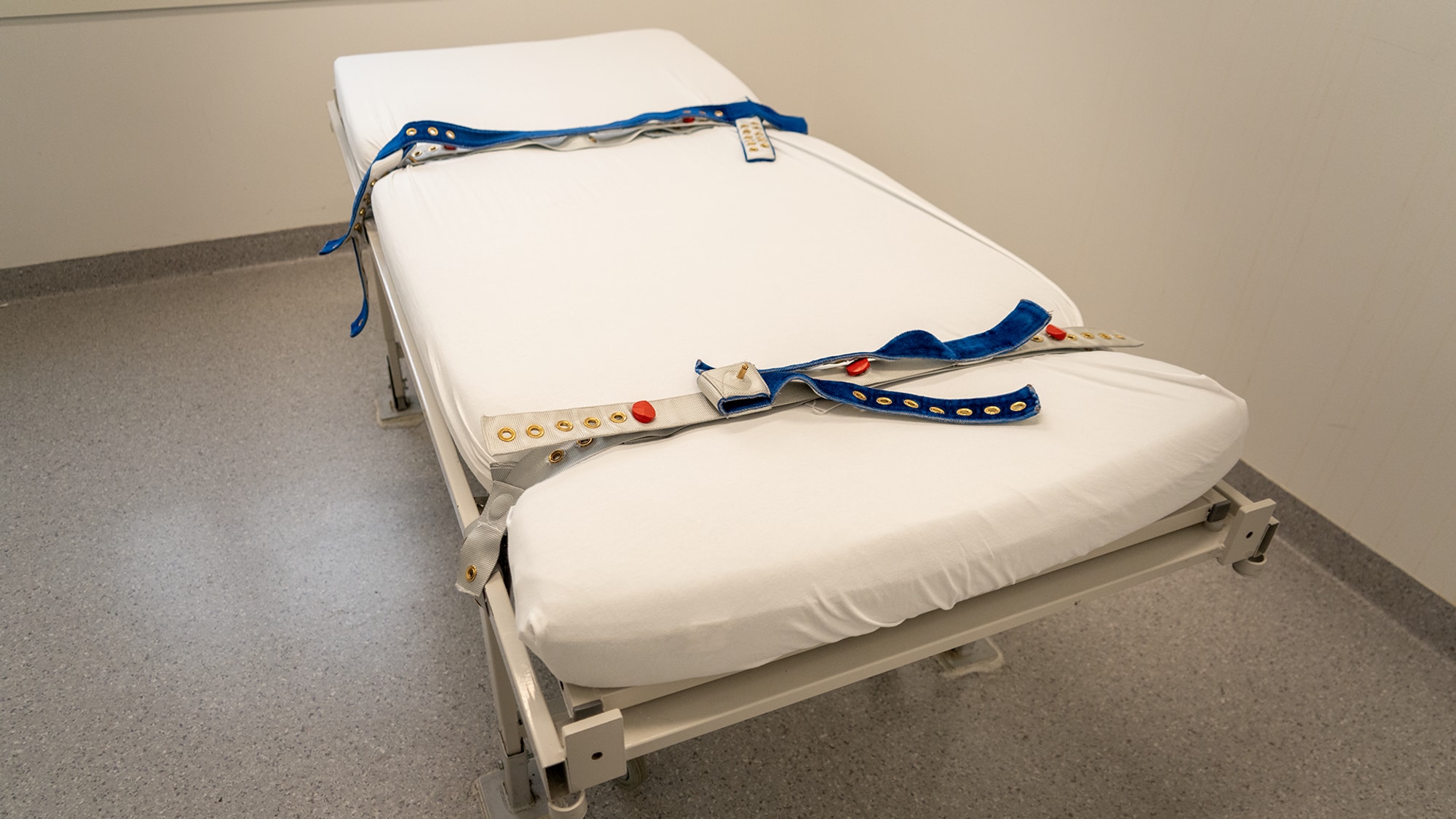 Et hvitt sykeseng med blå og grå stropper. Sengen er i et rom med lyse vegger og grå gulvbelegg. Stroppene er festet til sengen for å holde en person fast. (Bildebeskrivelsen er laget av en KI-tjeneste)