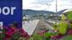 Synnøve Sundby Fallmyr Togstasjon med tog på skinnene og grønne fjell i bakgrunnen. En blå plakat med hvit tekst er delvis synlig i forgrunnen. Enkelte blomster er også synlige. (Bildebeskrivelsen er laget av en KI-tjeneste)