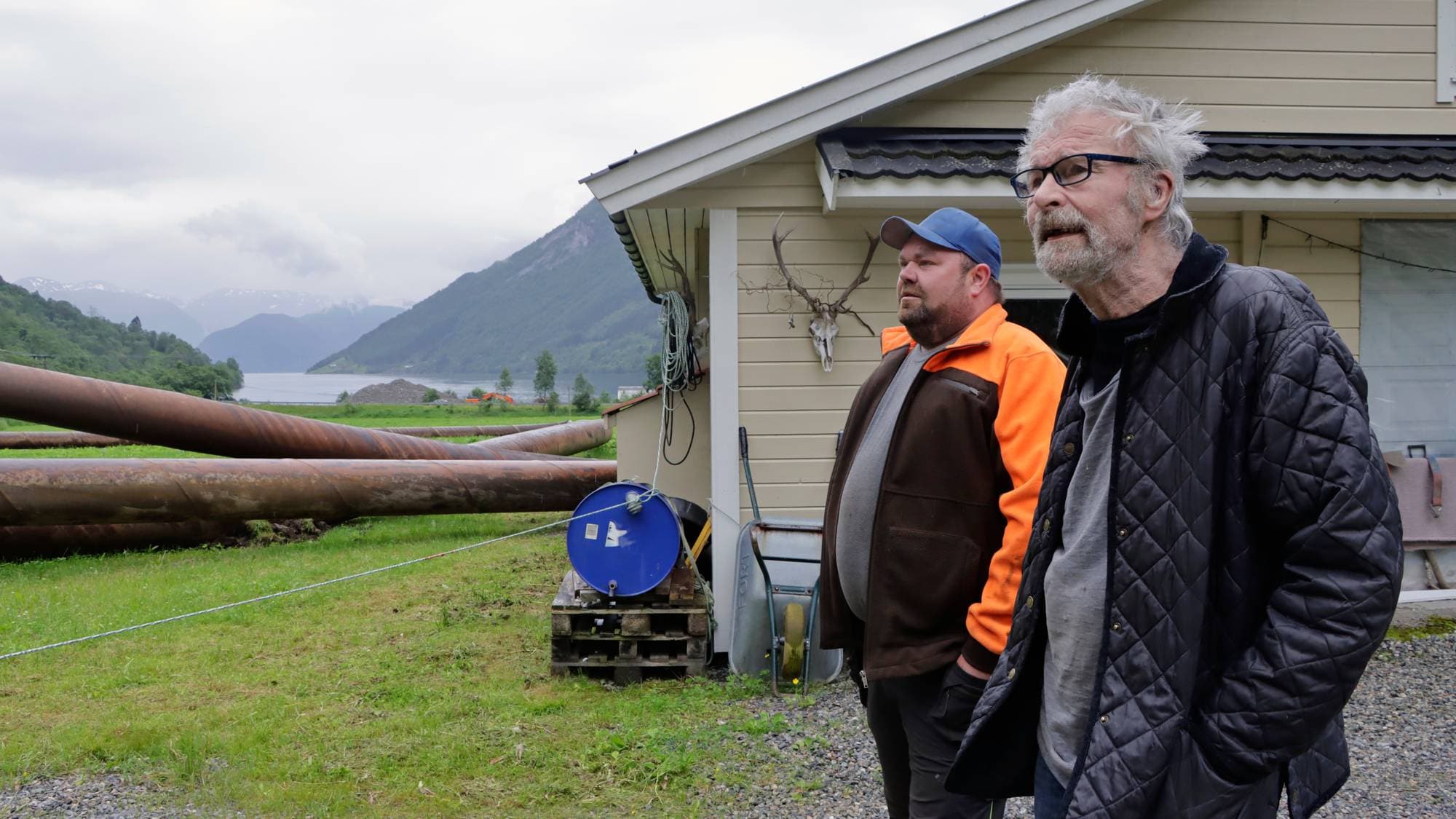 To menn står utendørs ved en bygning i et landskap med store rør og fjell i bakgrunnen. (Bildebeskrivelsen er laget av en KI-tjeneste)