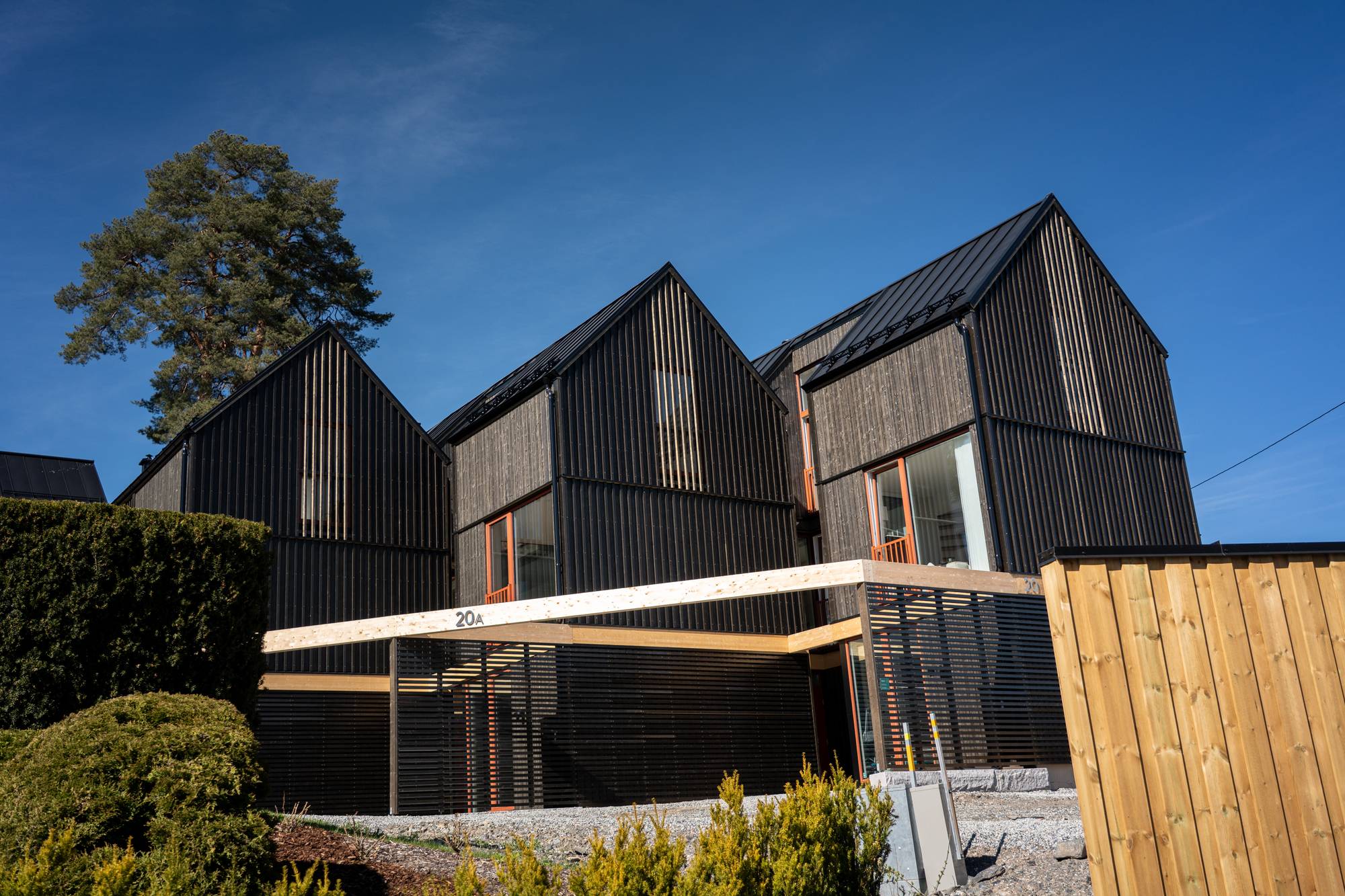 Moderne hus i tre under bygging med mørk fasade og store vinduer.
Bygget har spisse tak og utendørsareal med skjerming.
En stor furu står ved siden av bygget.
Det er en lys himmel og en trebarriere til høyre. (Bildebeskrivelsen er laget av en KI-tjeneste)