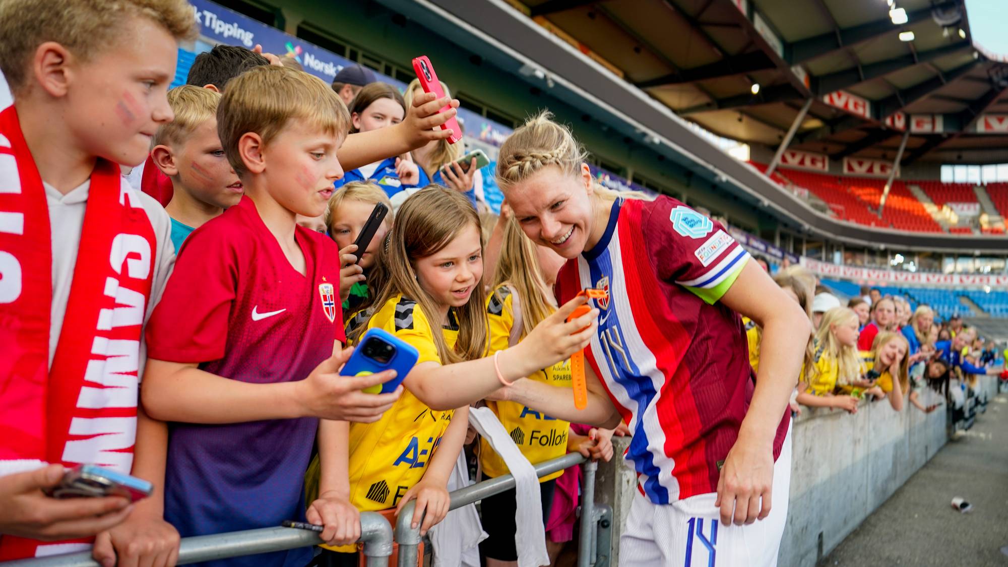 En fotballspiller signerer autografer til supportere på tribunen. De holder opp mobiltelefoner for å ta bilde. Spilleren er iført en rød, hvit og blå drakt. Supporterne er barn og ungdommer. (Bildebeskrivelsen er laget av en KI-tjeneste)