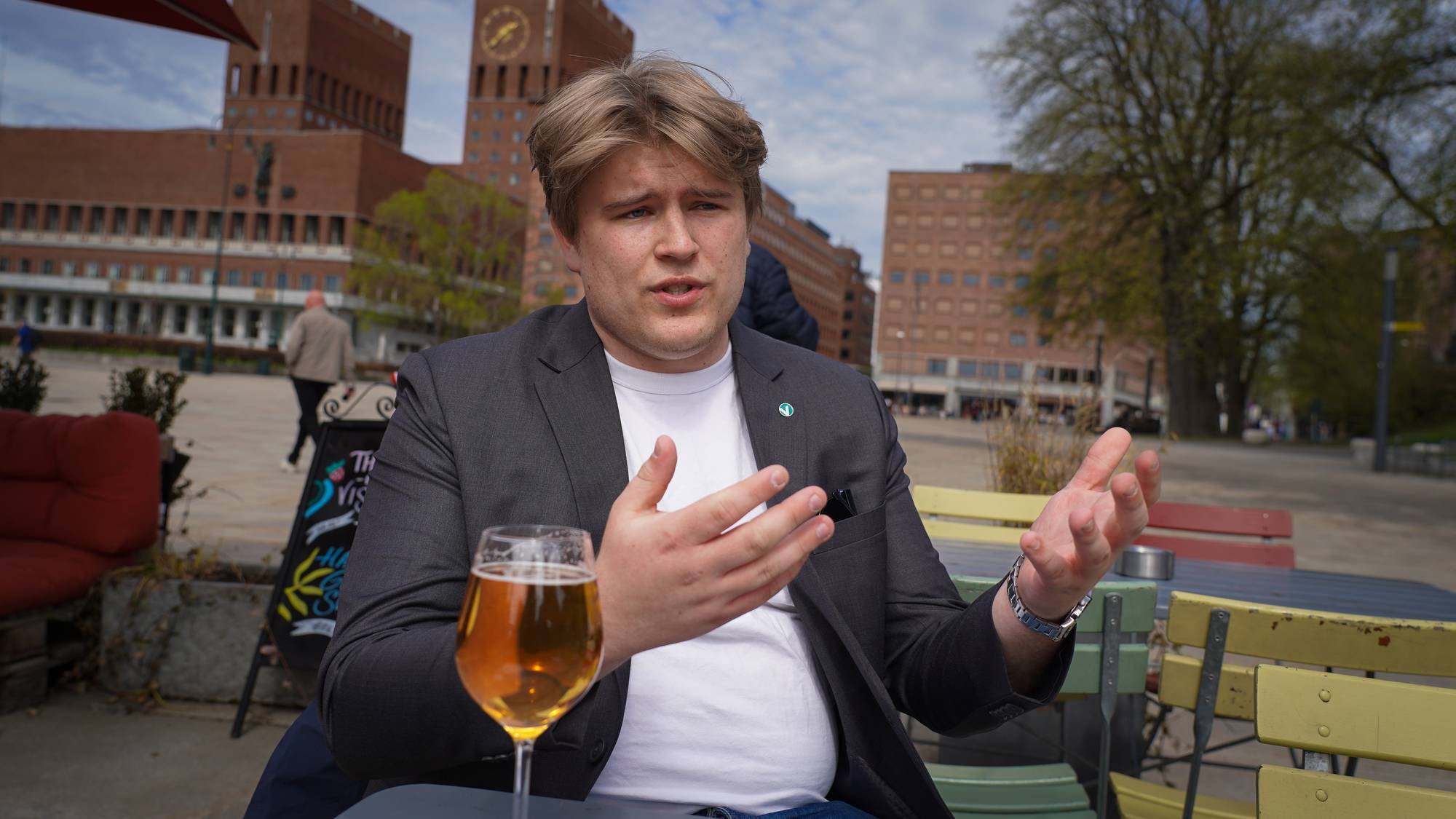 En ung mann i dressjakke snakker utendørs.
Han sitter ved et bord med et glass øl foran seg.
I bakgrunnen skimtes Rådhuset i Oslo.
Dette er et utendørs møte. (Bildebeskrivelsen er laget av en KI-tjeneste)