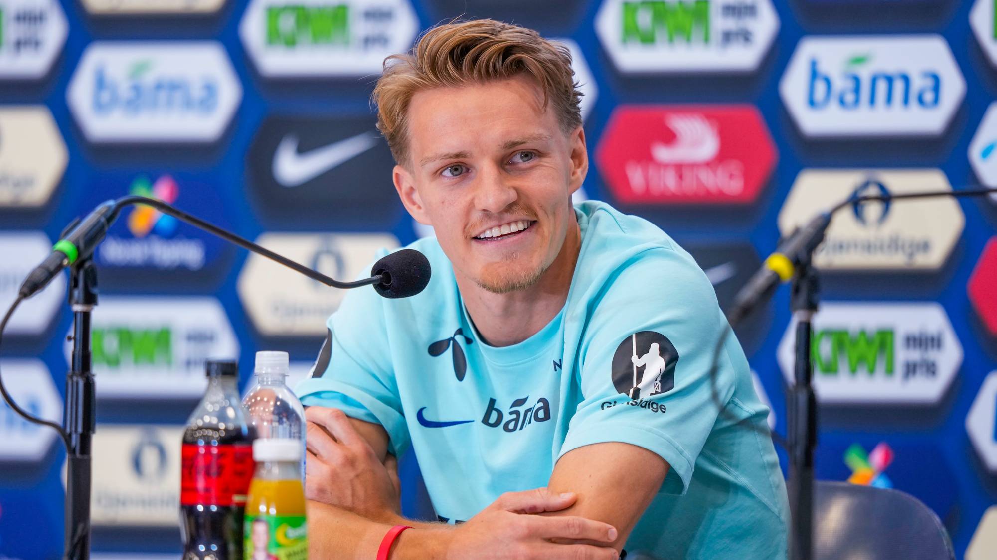 En mann sitter bak et bord med mikrofoner foran seg. Han smiler og ser mot siden. Mannen har på seg en lys blå t-skjorte. Bak ham er det en blå bakgrunn med flere logoer og sponsoravtaler. Foran mannen står det en flaske med mørk drikke, en flaske med vann og en liten flaske med oransje drikke. (Bildebeskrivelsen er laget av en KI-tjeneste)