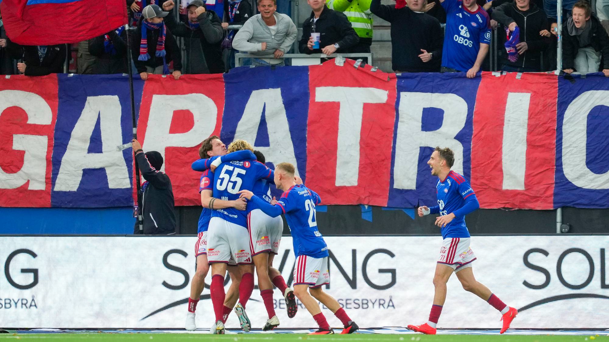 Fotballspillere i blå drakter feirer en scoring foran jublende supportere på tribunen. (Bildebeskrivelsen er laget av en KI-tjeneste)