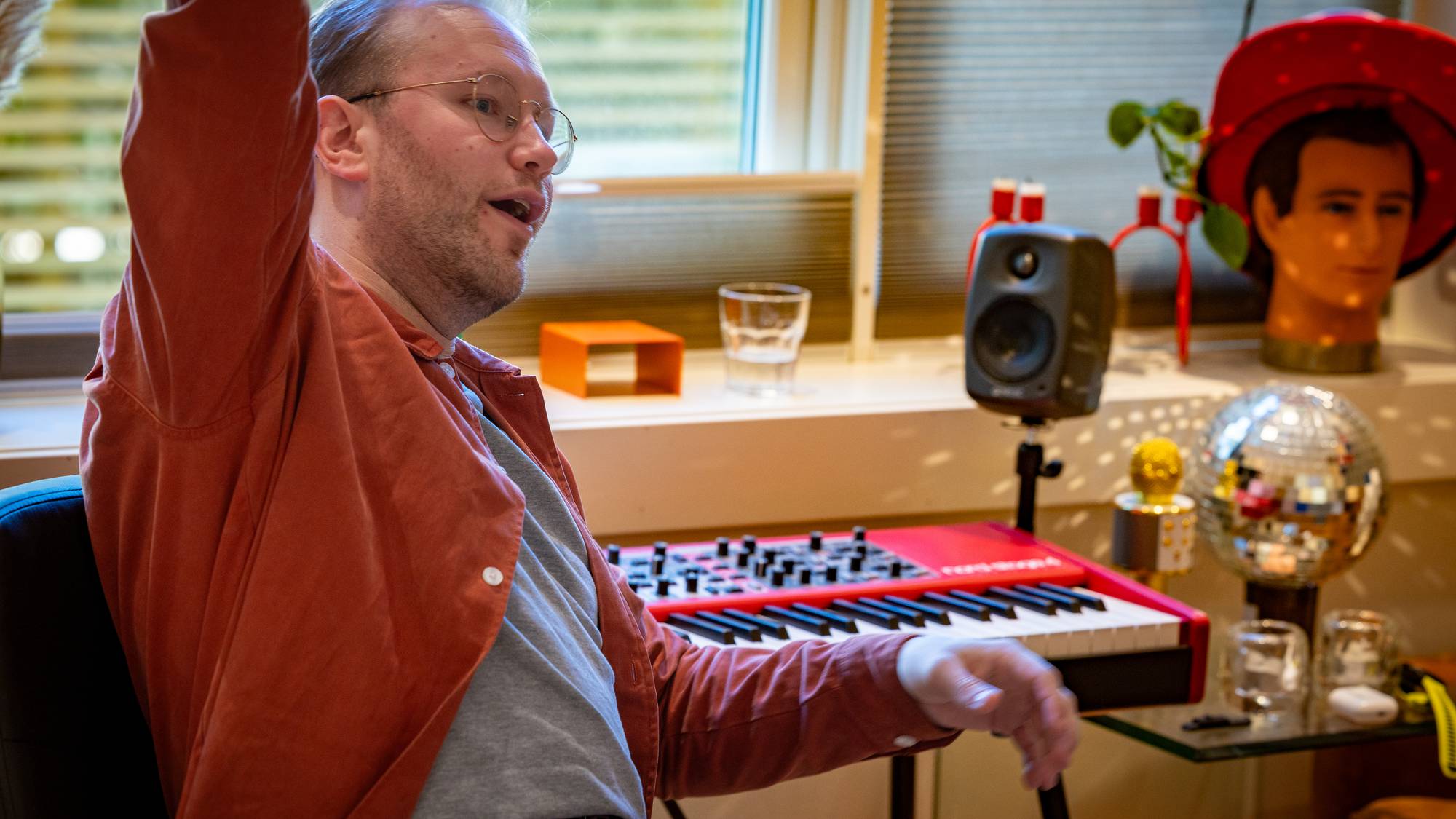 Mann med oransje skjorte løfter ein arm framfor eit raudt piano
