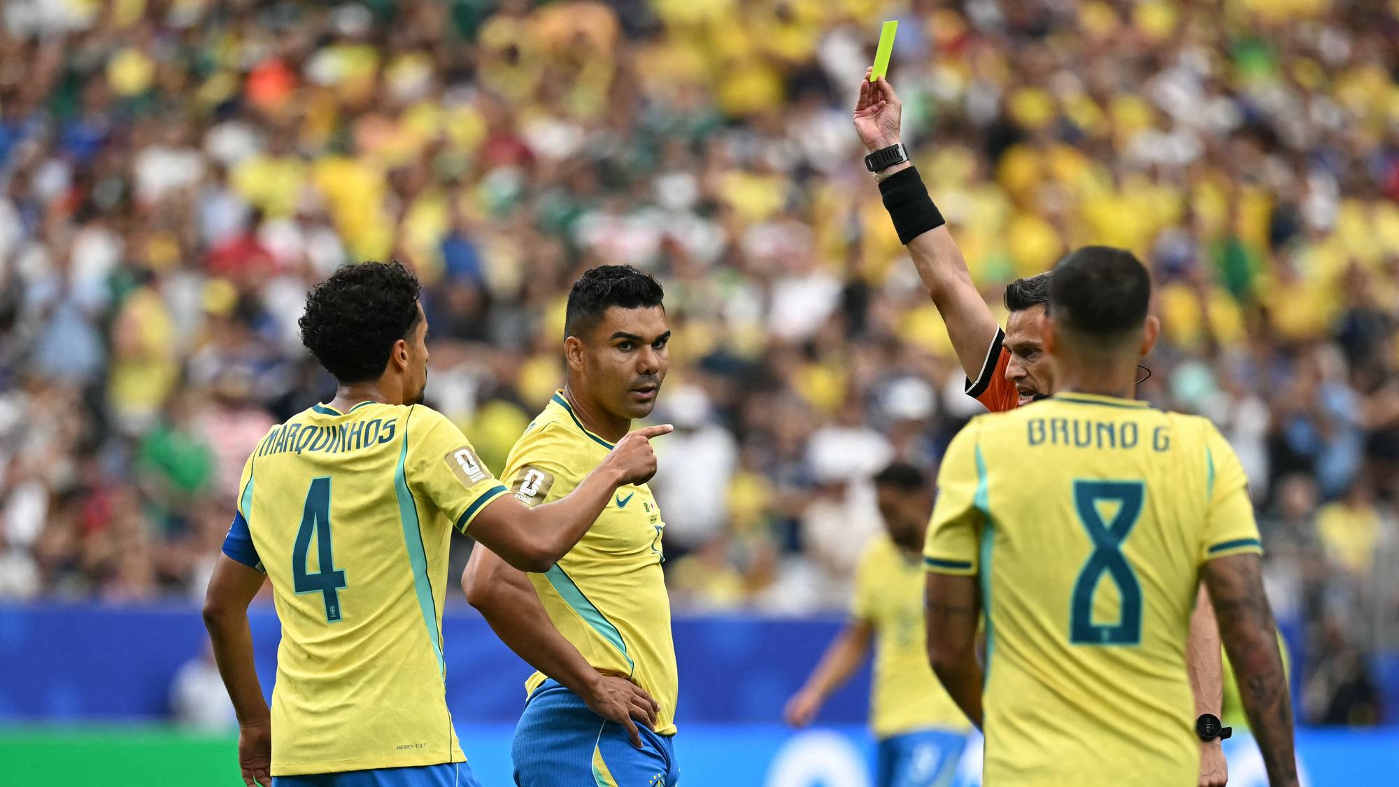 Dommer viser gult kort til en brasiliansk fotballspiller under en kamp foran et stort publikum. (Bildebeskrivelsen er laget av en KI-tjeneste)