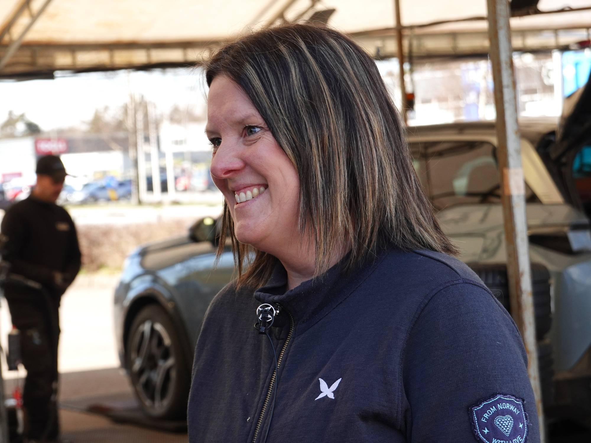 En kvinne med brunt og lyst hår smiler og ser til siden. Hun har på seg en mørk jakke med et hvitt symbol på brystet. Bak henne står en mann i en svart caps og jakke, med en bil i bakgrunnen. De befinner seg utendørs under en takkonstruksjon. (Bildebeskrivelsen er laget av en KI-tjeneste)