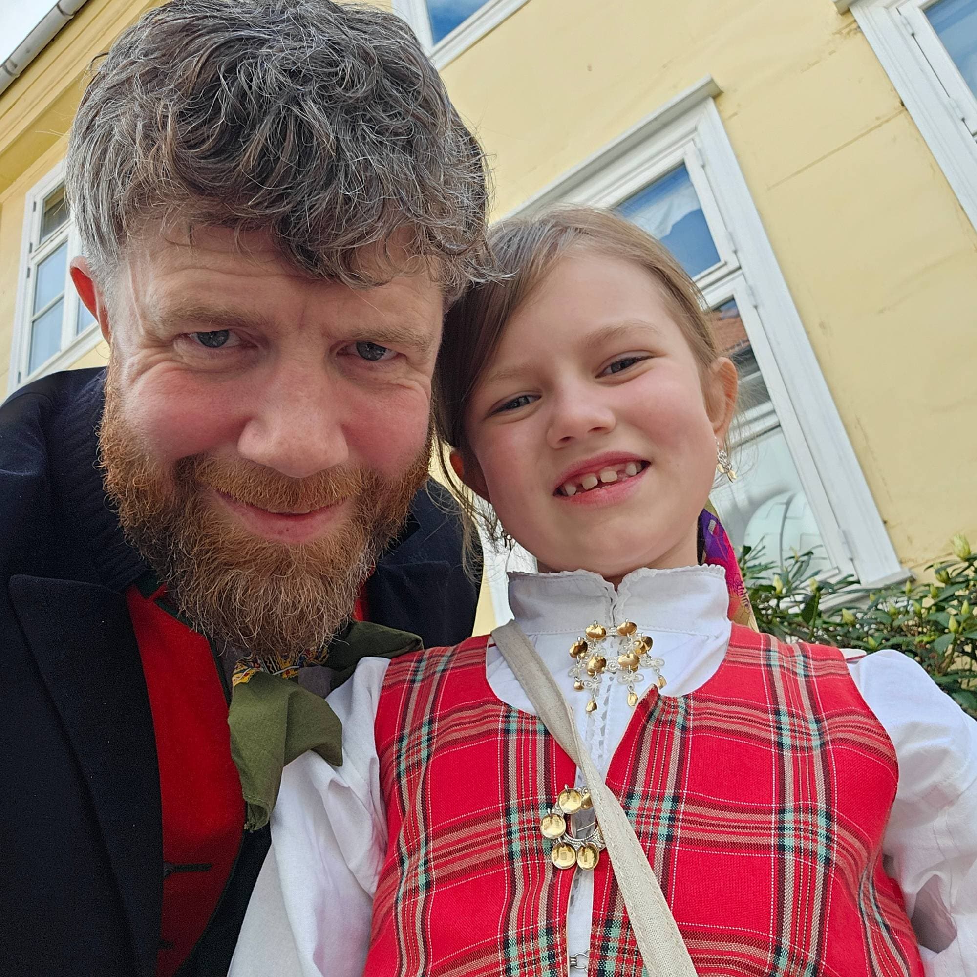 En mann med skjegg og en jente ser inn i kamera. Bak dem er det et gult hus. Mannen har på seg en svart bunad med rødt og grønt i halsen. Jenta har en festdrakt med hvit skjorte og rødrutete vest. 