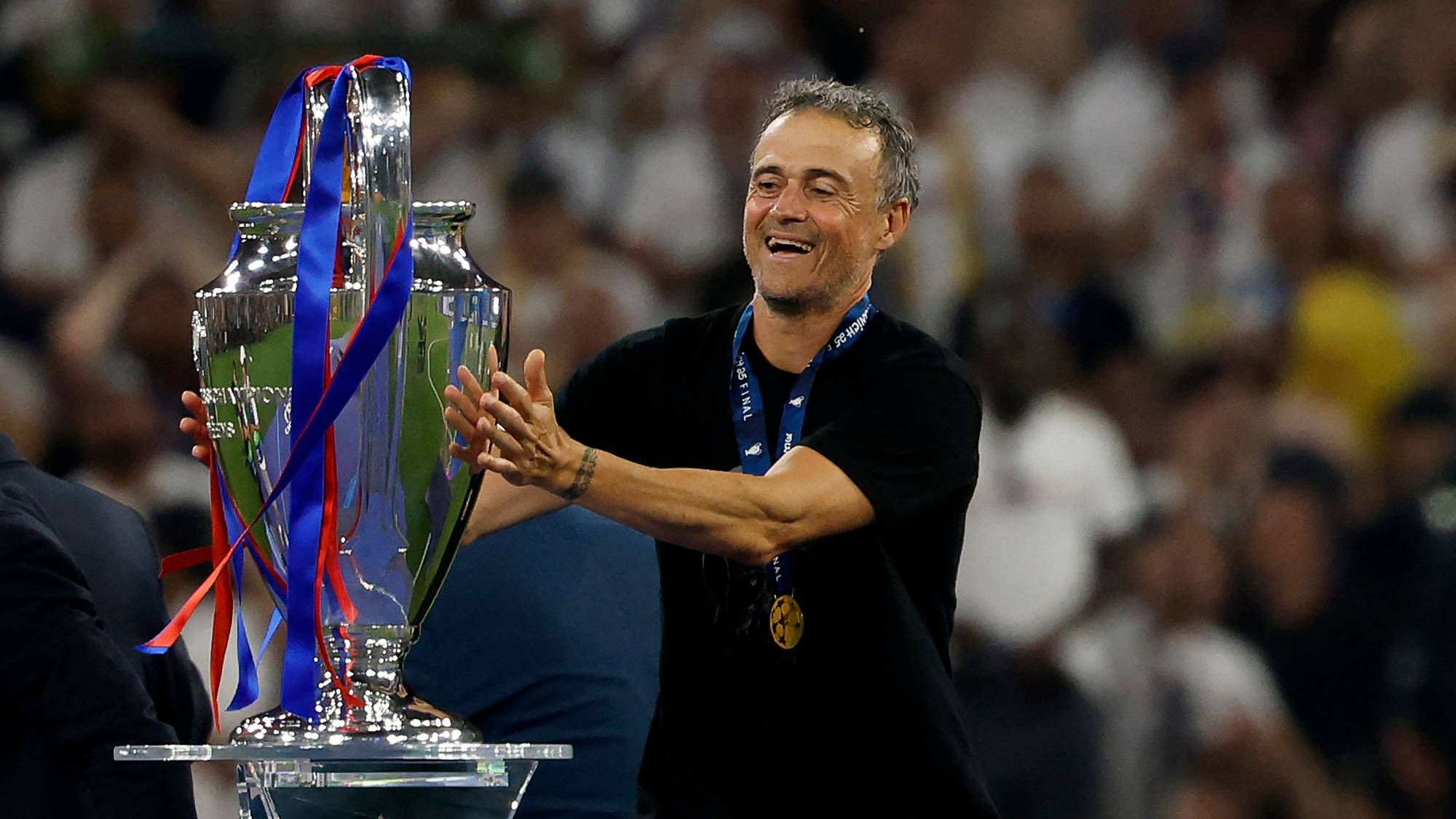 En mann med mørk t-skjorte holder Champions League-trofeet. Han ler og har medalje rundt halsen. I bakgrunnen sitter mange mennesker på tribunen. (Bildebeskrivelsen er laget av en KI-tjeneste)
