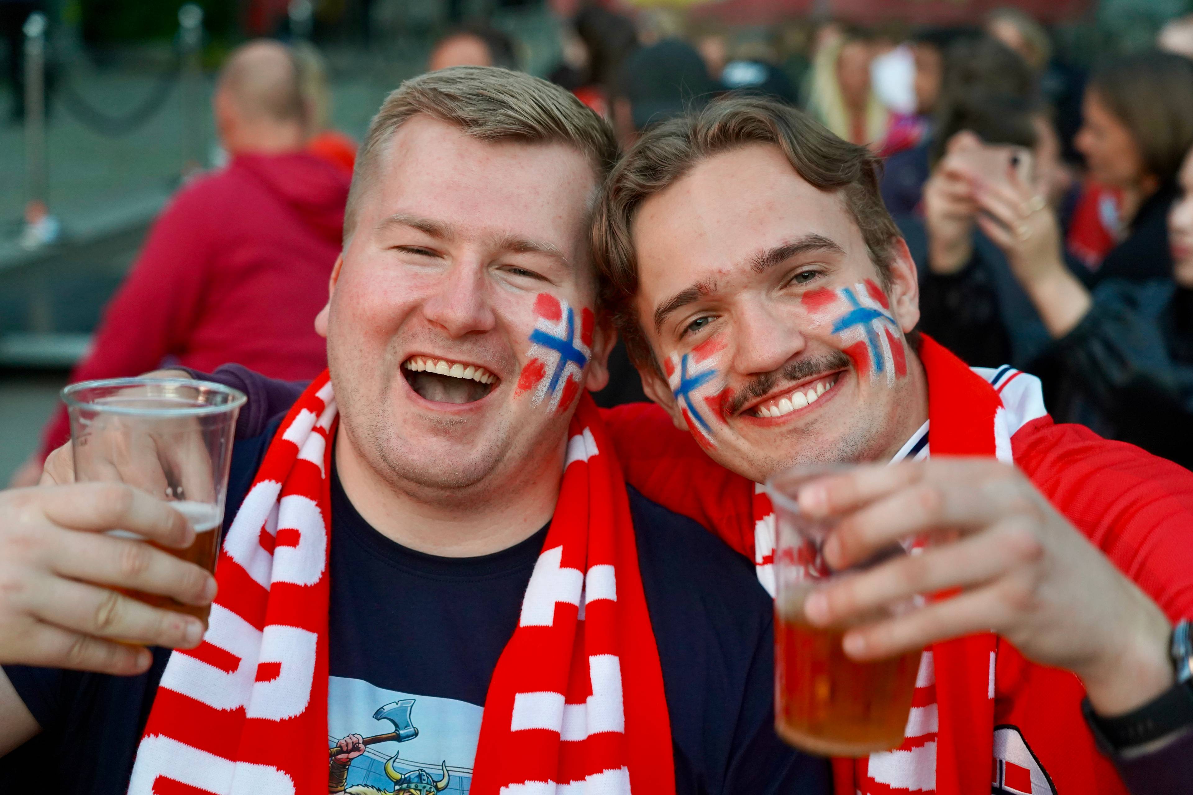 To menn med norsk flagg malt i ansiktet og røde og hvite skjerf smiler til kamera. De holder hvert sitt glass med øl. Bak dem står en folkemengde.